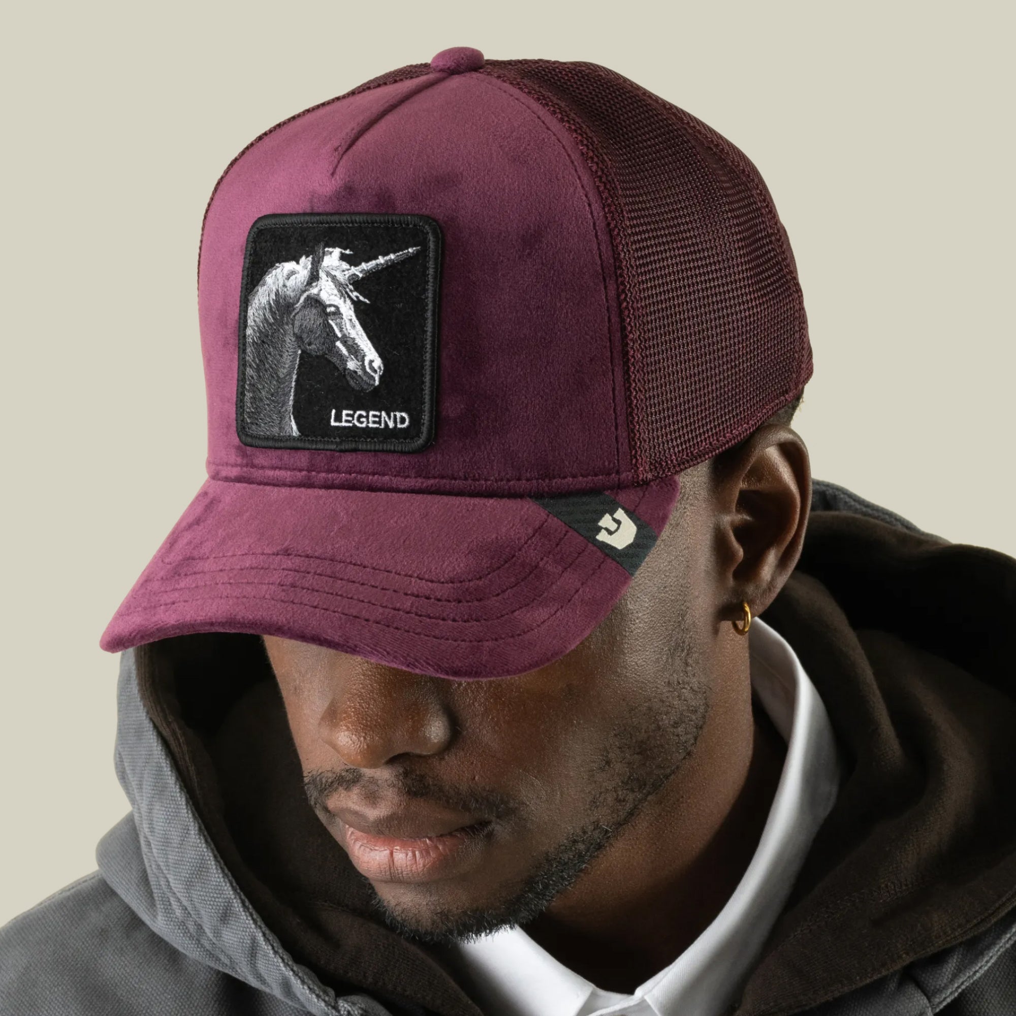 A man wearing the Goorin Bros. Velour Legend, a burgundy velour trucker hat with a breathable mesh back and a monochrome unicorn patch reading “Legend” on the front, looks slightly downward while dressed in a grey hoodie and white shirt.