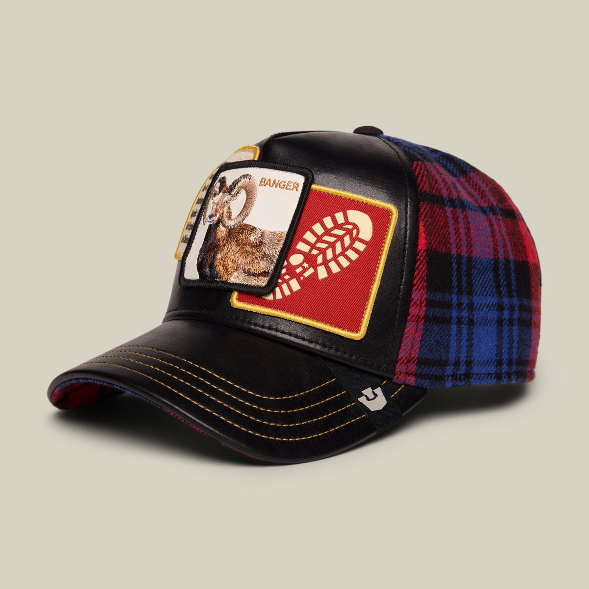 The Goorin Bros. Banger trucker hat features a black front panel with embroidered animal patch and red and gold logo, paired with a red and navy plaid fabric back and side panels, displayed against a neutral studio background.