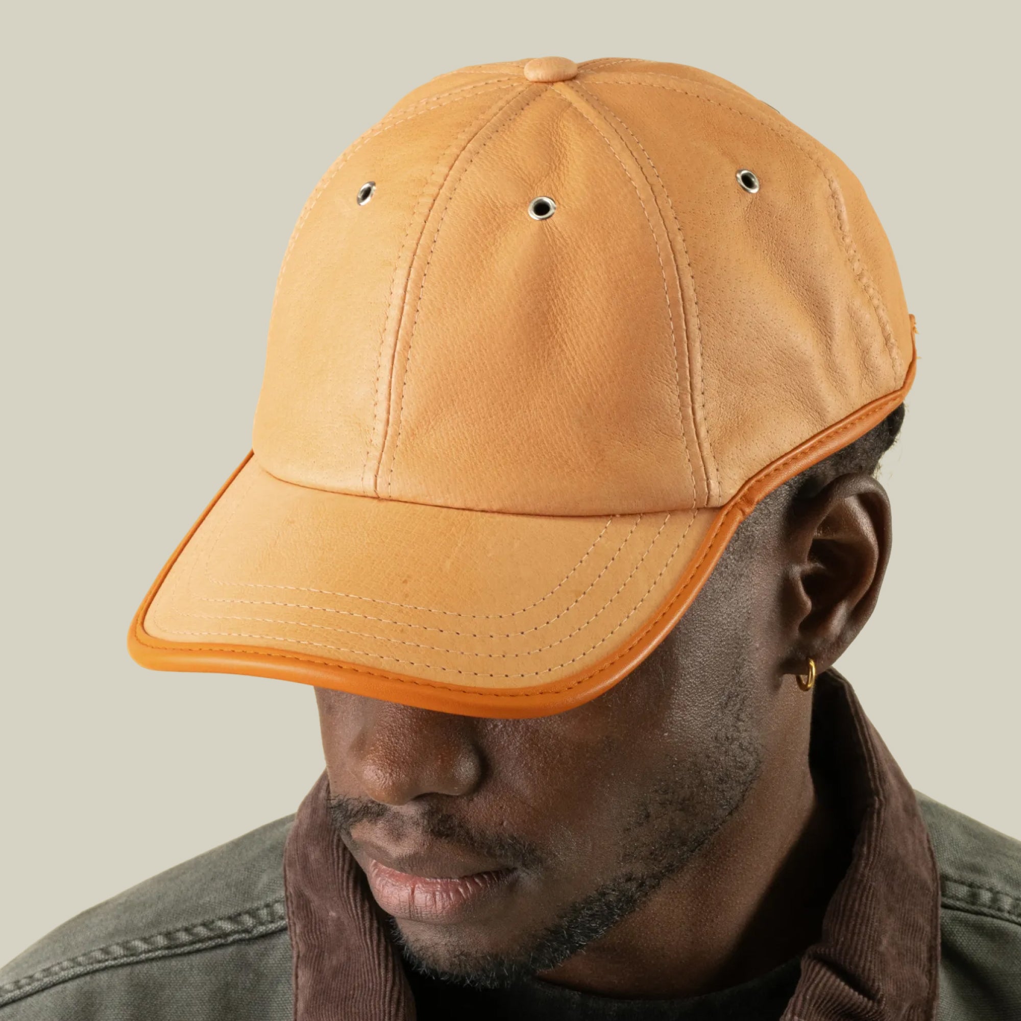 A person wearing the Goorin Bros. Steiner tan suede papa cap with orange trim and an adjustable plaid strap, looking down against a plain light beige background.