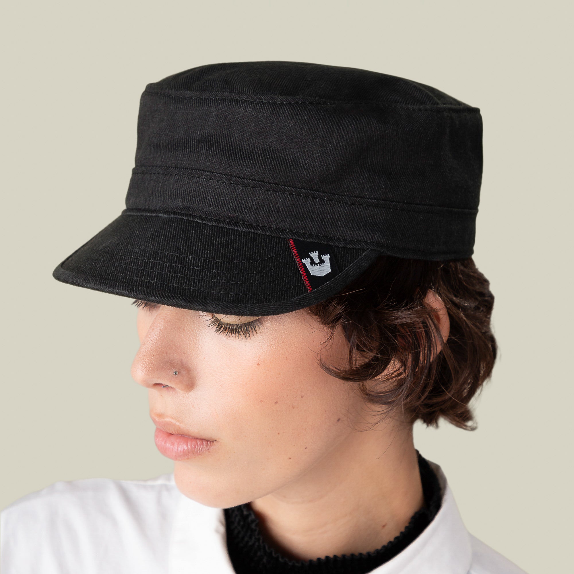 A person in a white top looks downward, wearing the Goorin Bros. Home at Last hat—a stylish, lightweight black cadet cap with a small crown logo patch—against a plain background.