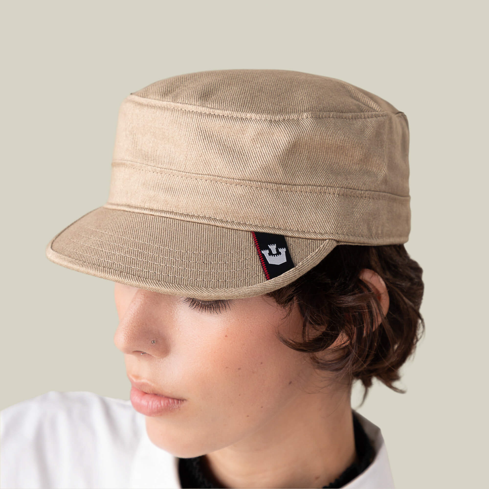 A person wearing the Goorin Bros. "Home at Last" beige military-style cap, featuring a small black and red logo tag, looks downward against a plain light background.