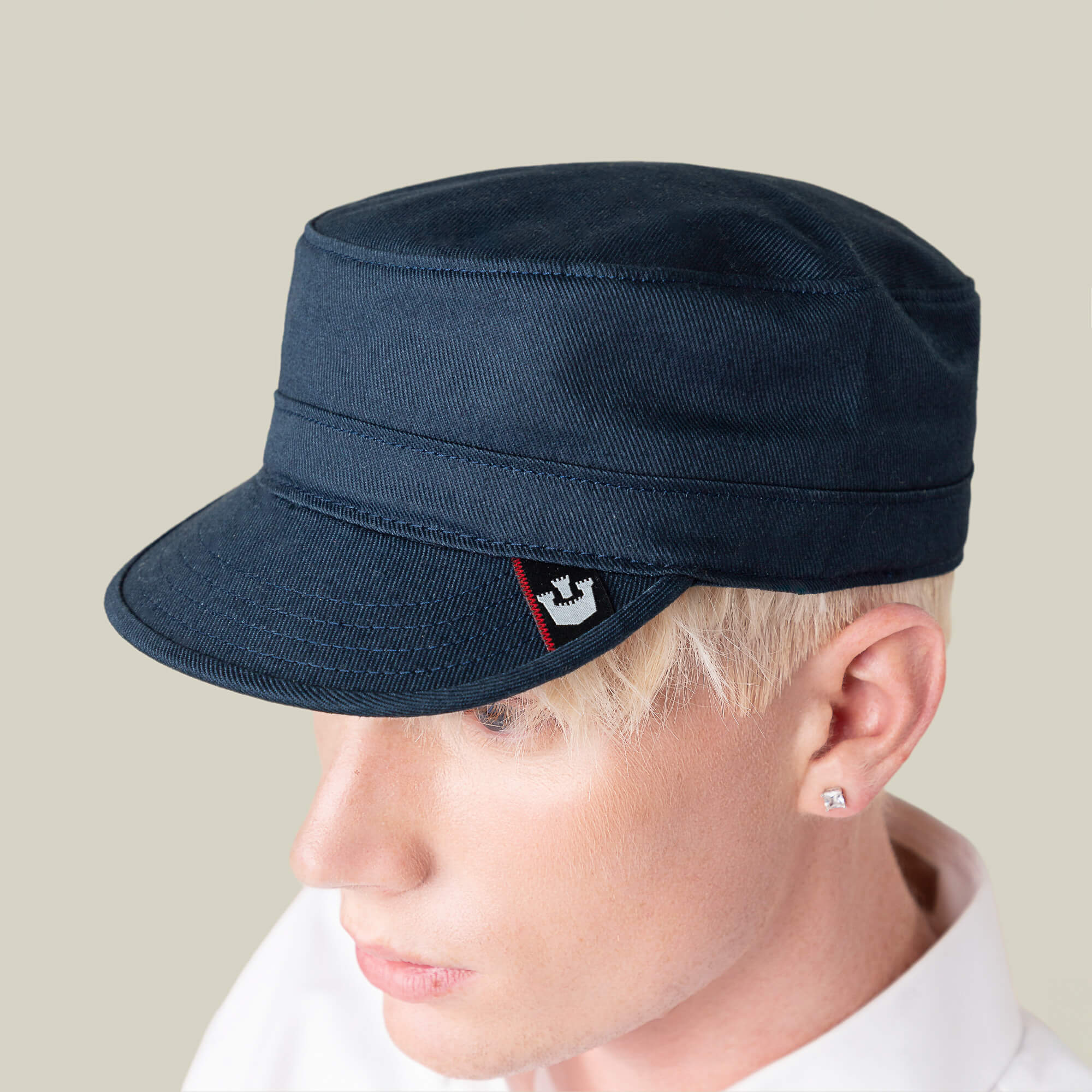 A person wears the Goorin Bros. "Home at Last" navy military-style cap with a triangular patch, paired with a white collared shirt, set against a plain beige background.