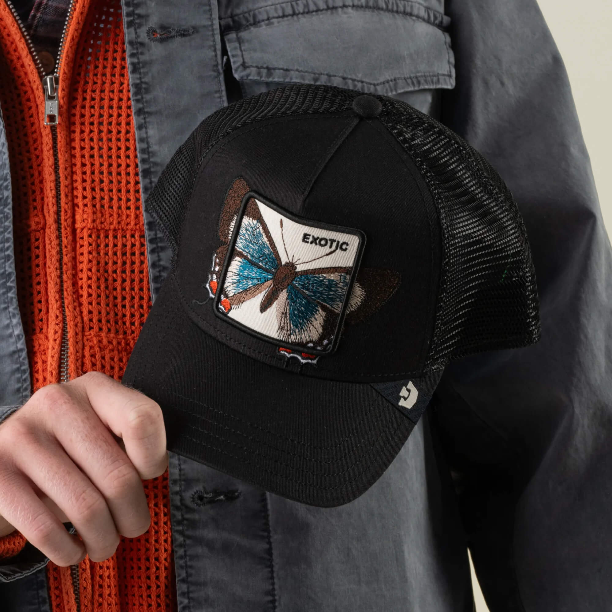 A person holds the Goorin Bros. Over the Line Butterfly cap, a black breathable mesh hat featuring a butterfly patch with “EXOTIC” embroidered above it. The butterfly-embroidered accessory complements their outfit, paired with an orange zip sweater and a gray jacket.