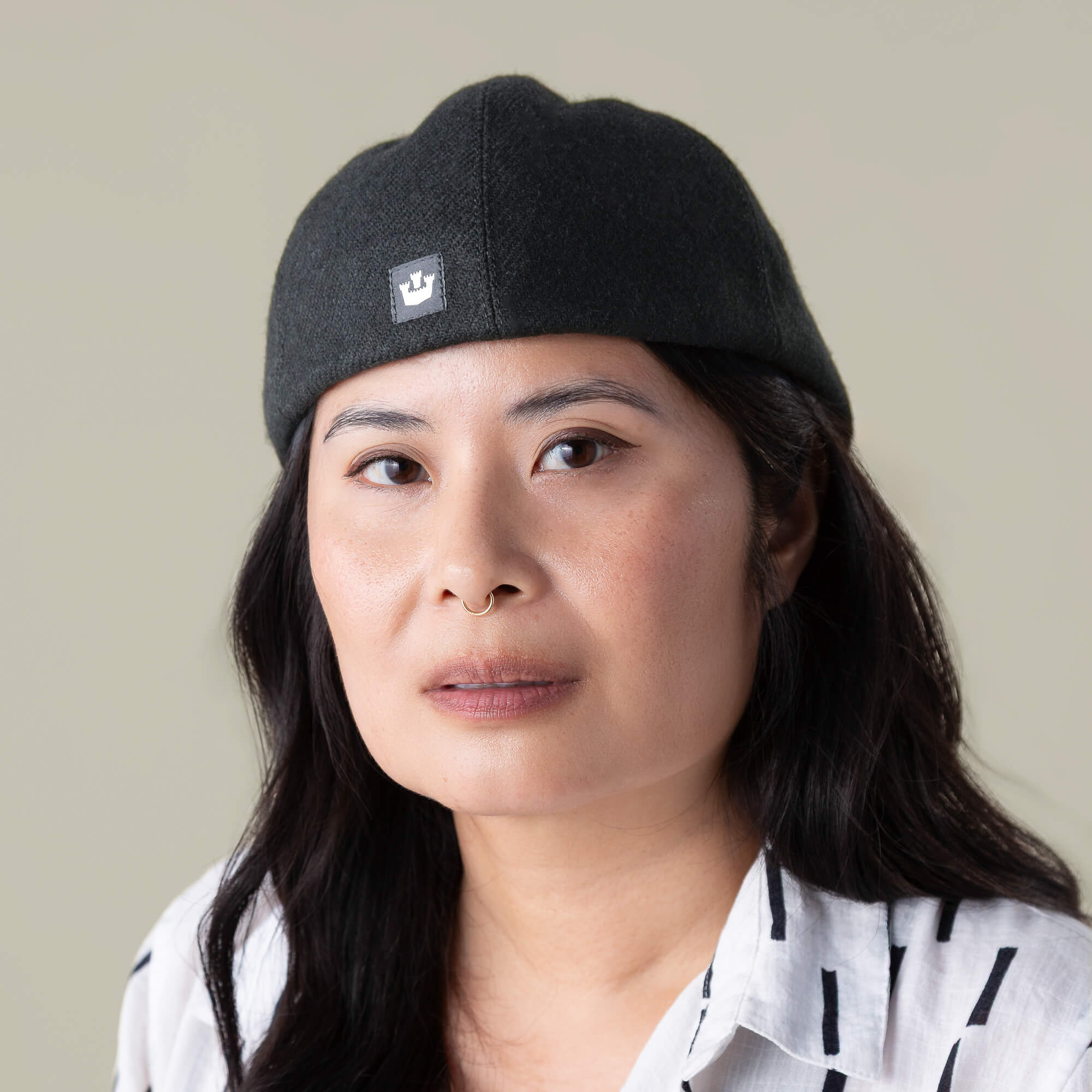 A person with long dark hair wears the Ronald Amos everyday cap by Goorin Bros., featuring a two-inch brim, paired with a white shirt with black stripes, and looks at the camera with a neutral expression.