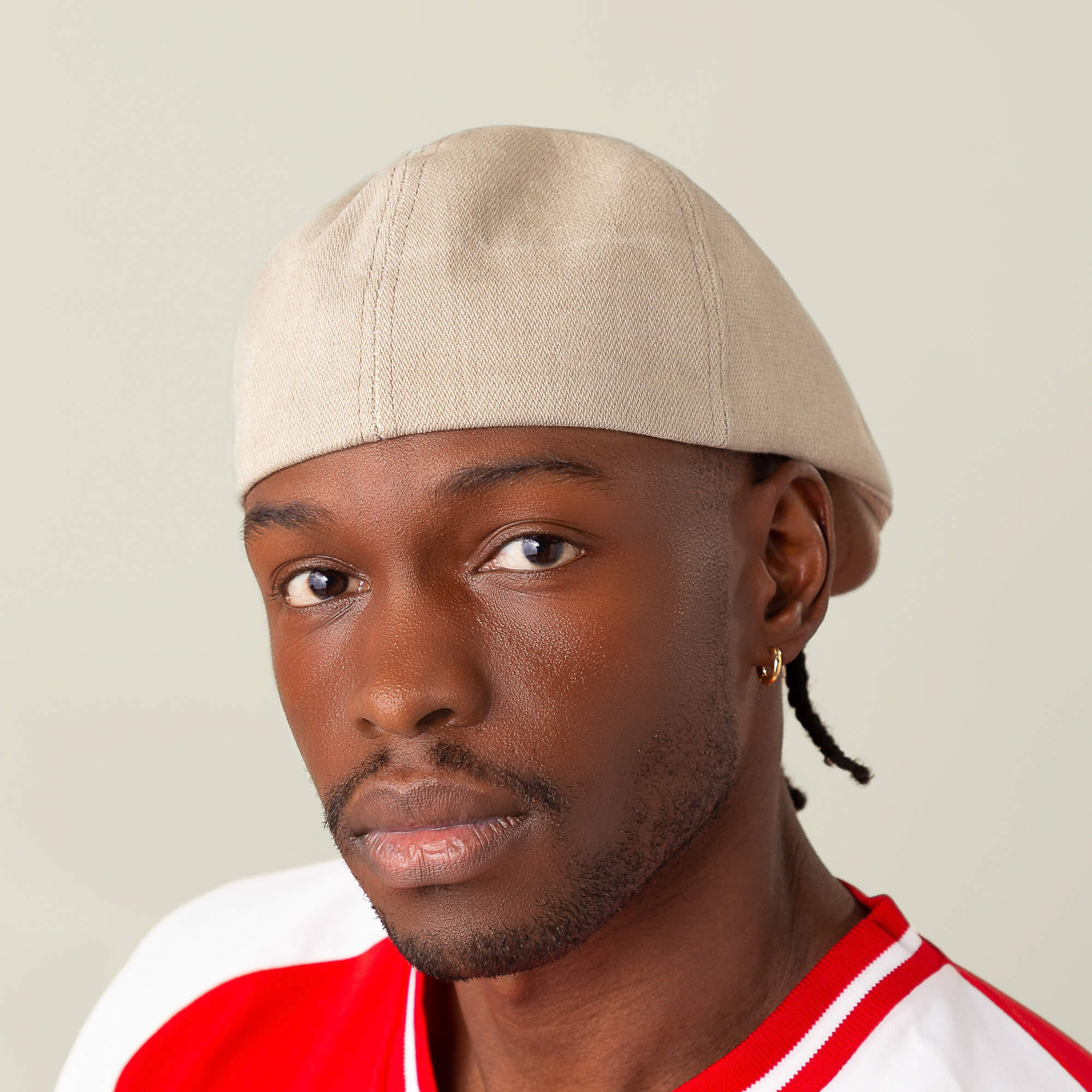 A man wears the Noisy Benjamin Paul beige cap by Goorin Bros., along with a red and white shirt and a gold hoop earring, looking directly at the camera against a plain light background.