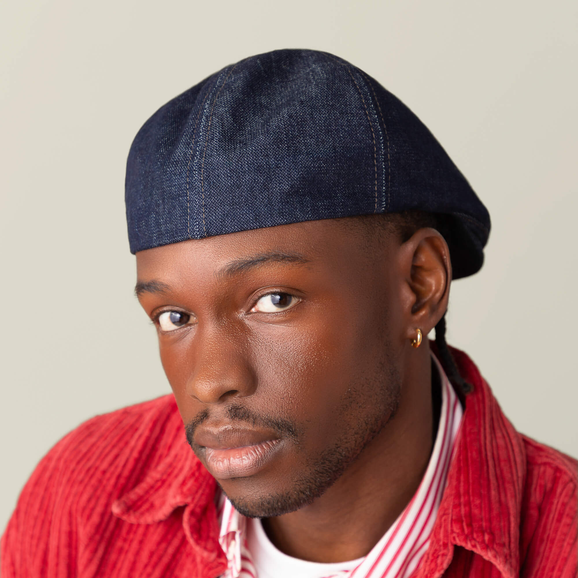 A person wears the Goorin Bros. Noisy Benjamin Paul navy flatcap, gold hoop earring, red corduroy jacket, and striped shirt while looking directly at the camera against a plain background.