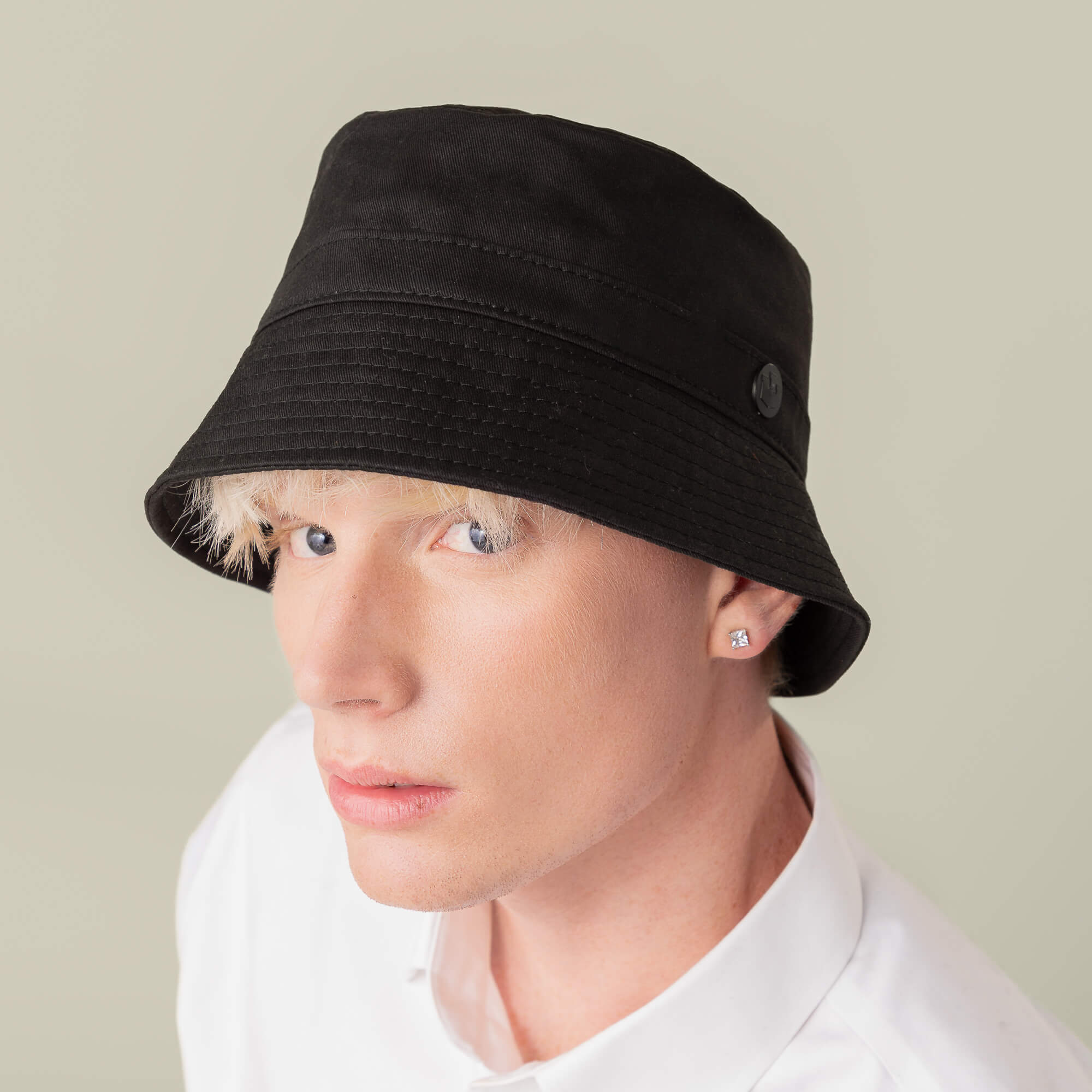 Person with light skin and short blond hair wears the Goorin Bros. Odyssey Bucket hat in solid black cotton, paired with a white collared shirt and stud earrings, looking at the camera against a plain background.