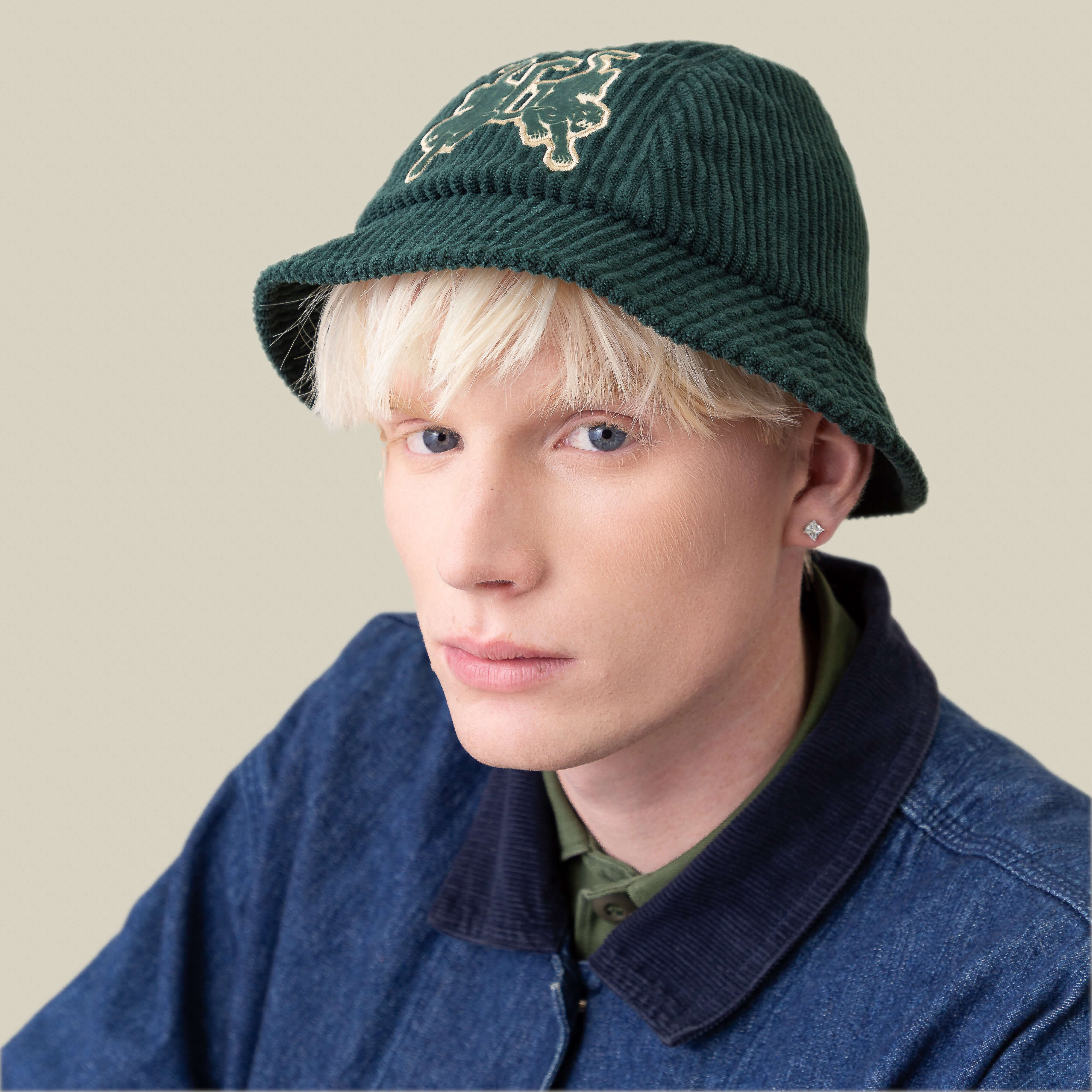 A person with blond hair wears the Goorin Bros. Yes Coach green corduroy bucket hat with embroidered design, a dark jacket, and a green collared shirt, seated against a plain light background.