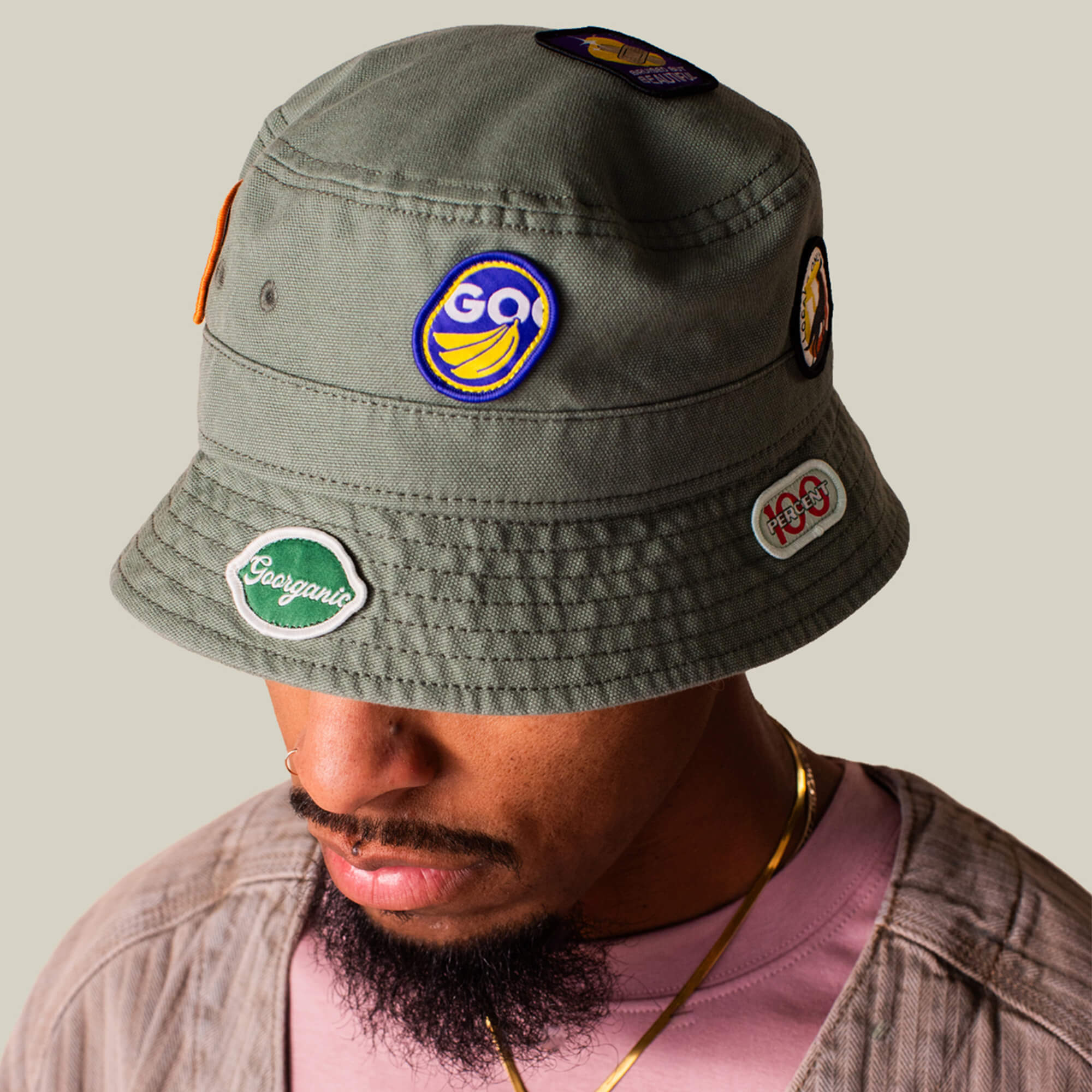 A man wearing the Goorin Bros. PLU 1895 olive green bucket hat, featuring colorful banana and text patches, looks down against a neutral background.