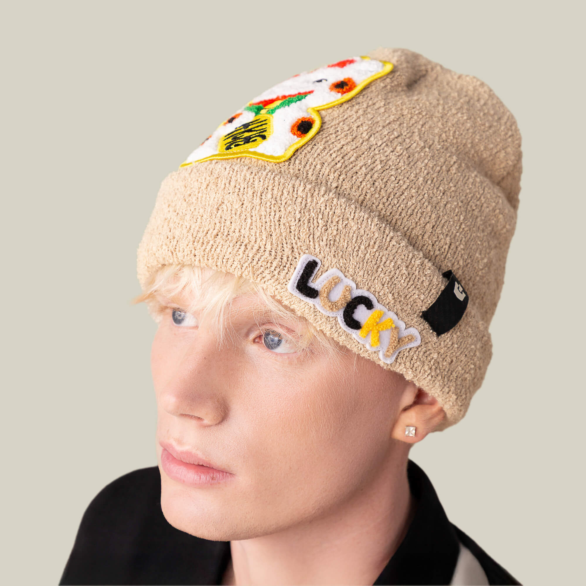 A light-skinned, blond-haired person wears a cozy tan "Good Fortune" beanie by Goorin Bros., featuring patch designs and a warm knit texture, looking slightly left against a plain background.