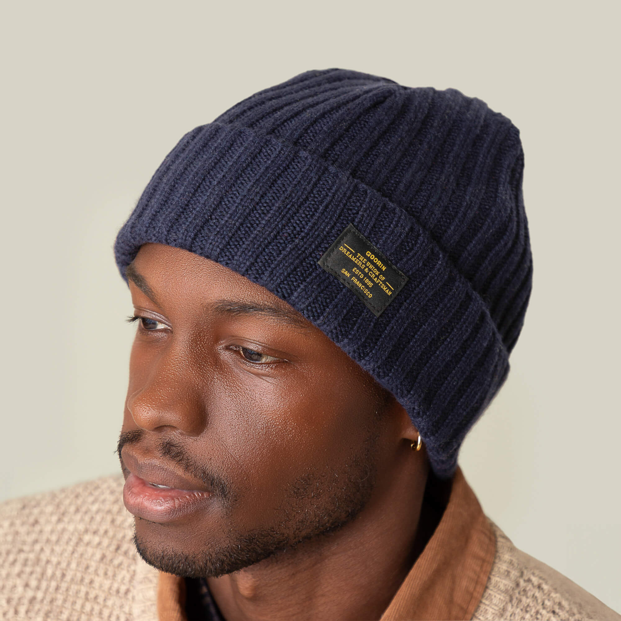 A man wears the navy blue Dream Cap by Goorin Bros.—a luxurious cashmere wool knitted beanie with a black label—paired with a beige textured jacket and brown shirt against a neutral background.