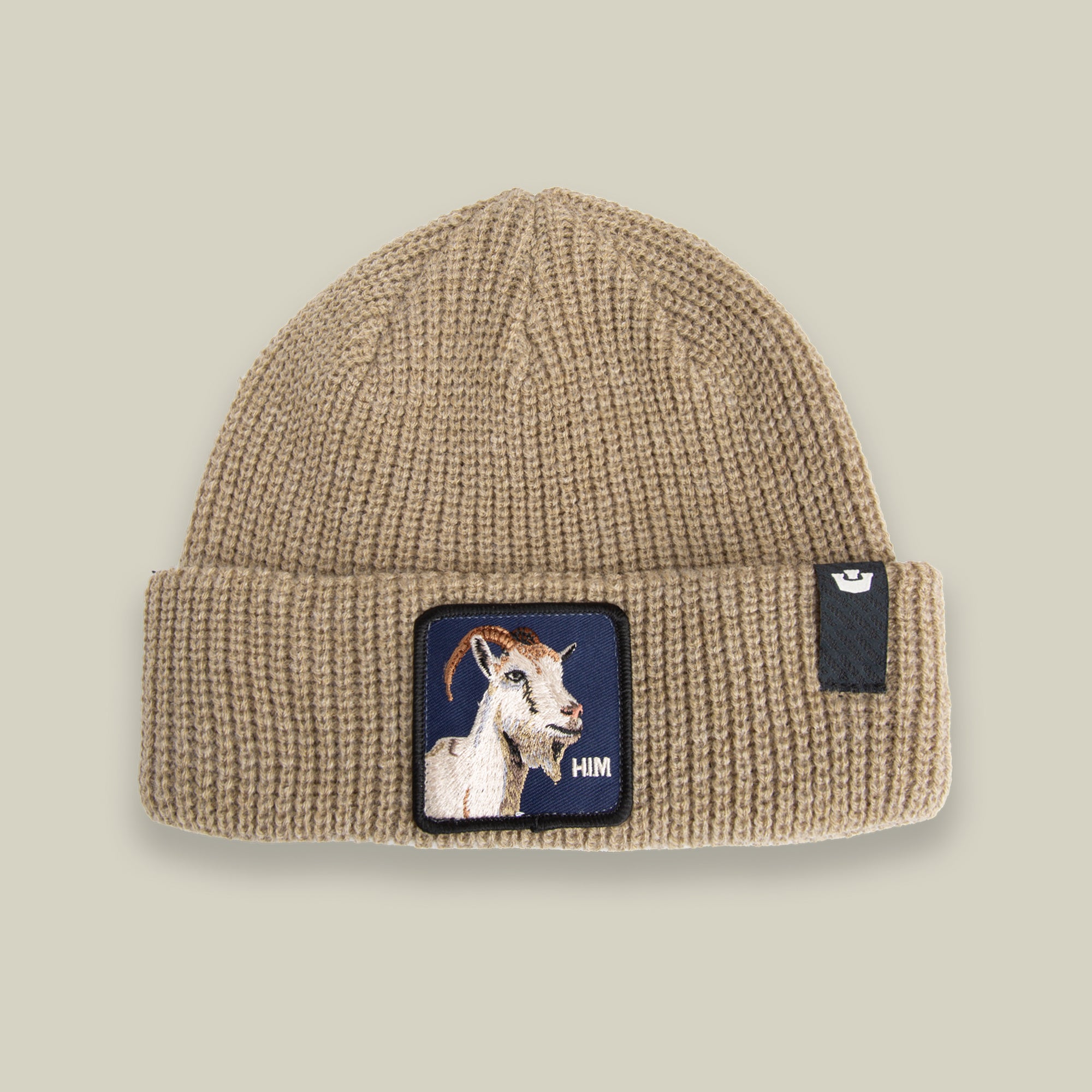 Stay warm in The Him Beanie by Goorin Bros.—a BISCUIT-colored knit with a cuff, bold goat patch, and "HIM" text on the front, plus a black tag with a white logo on the right side.