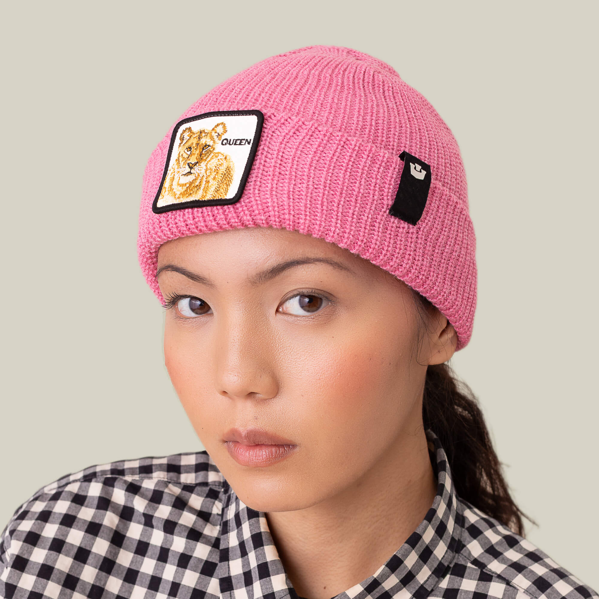 A person wears Goorin Bros.' The Queen Beanie, a pink knit winter hat with a lioness patch and QUEEN text, paired with a black-and-white checkered shirt against a plain background.