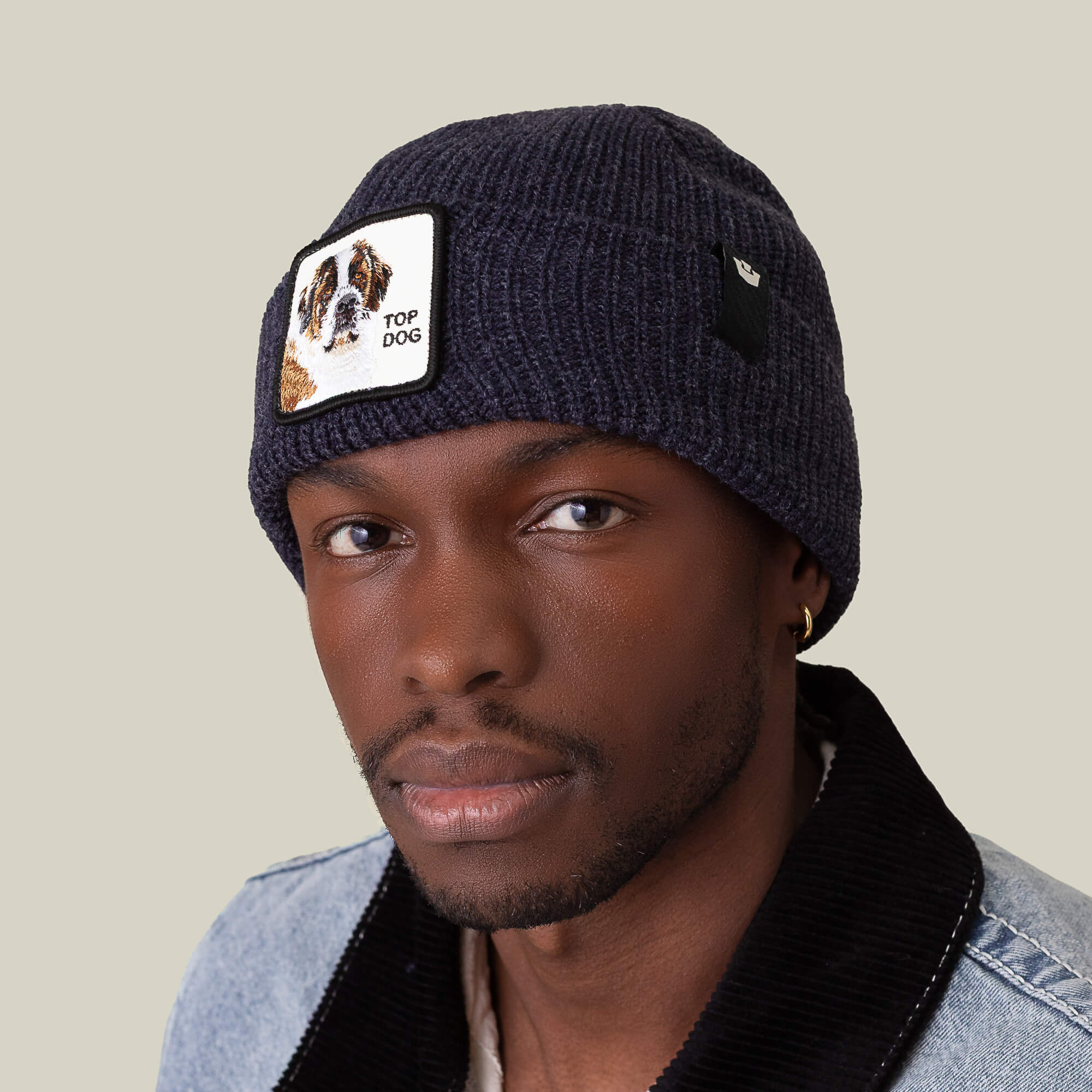 A man wears The Top Beanie by Goorin Bros., a navy knit hat featuring a St. Bernard dog patch, paired with a denim jacket with a black collar.