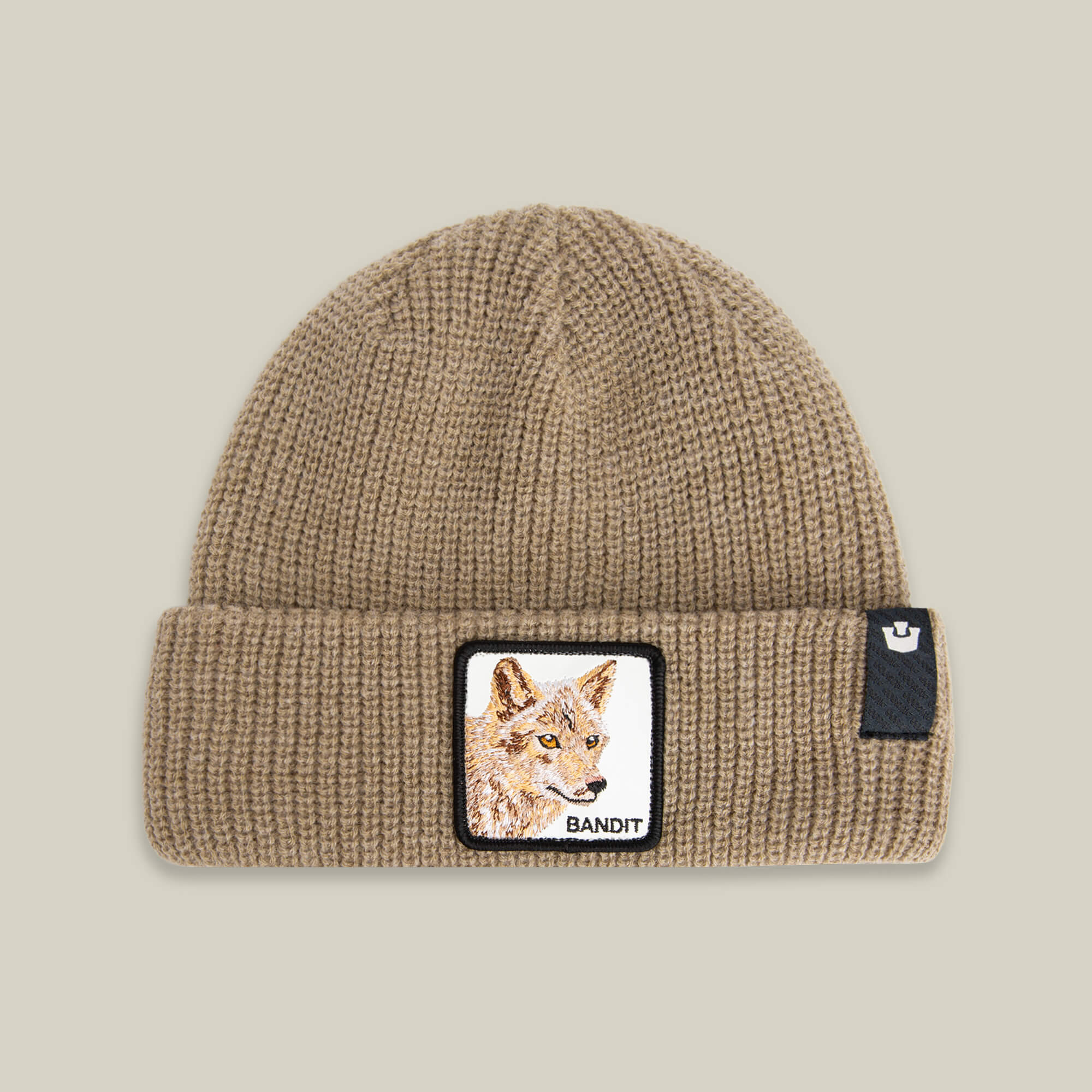 The Bandit Beanie by Goorin Bros. in BISCUIT features an earth-tone knit, a front patch with a wolf labeled BANDIT, and a black tag on the cuff—making it an ideal, cozy winter hat.