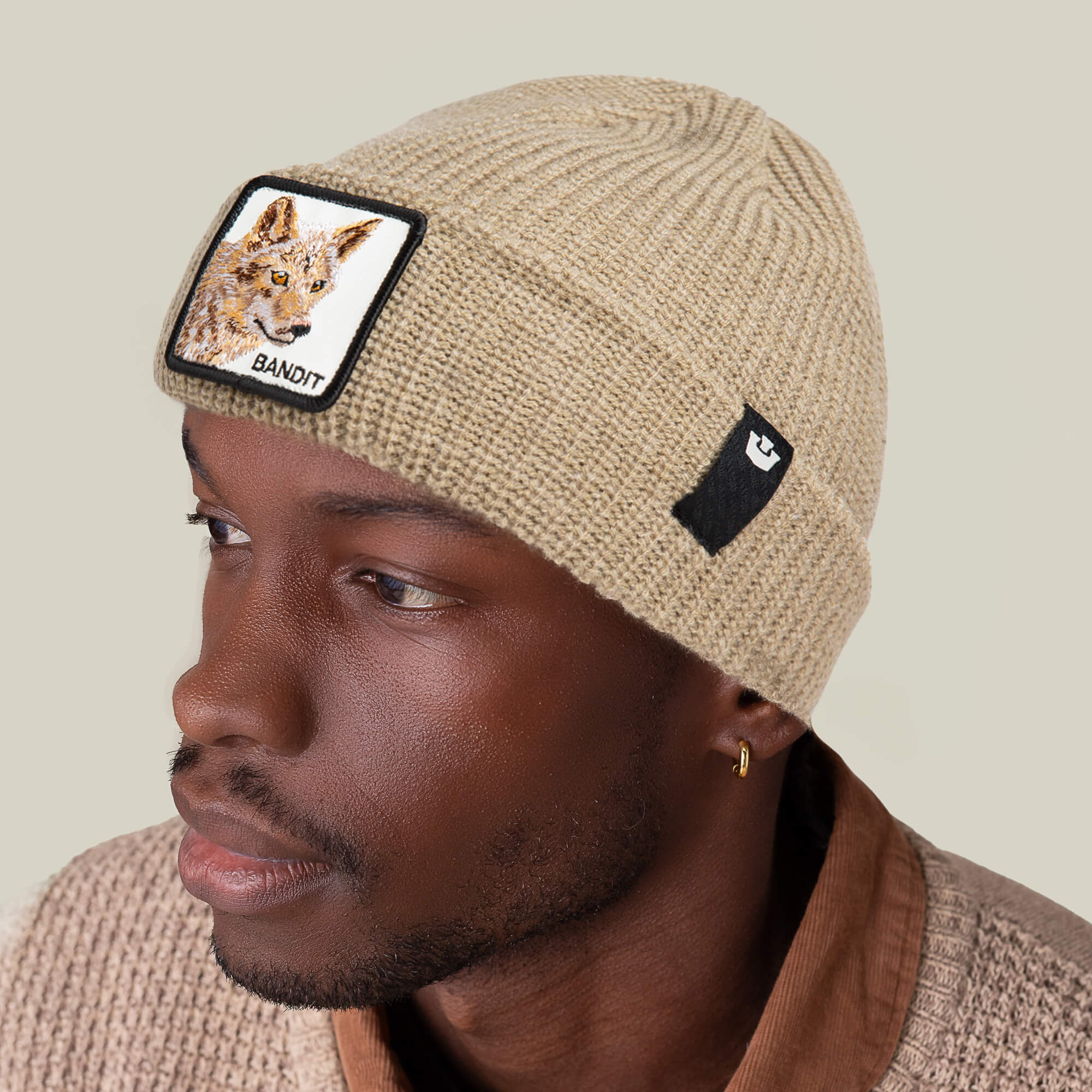 A man looks off to the side wearing The Bandit Beanie by Goorin Bros., an earth-tone knit winter hat featuring a coyote patch with the word "BANDIT" and a small black logo patch.