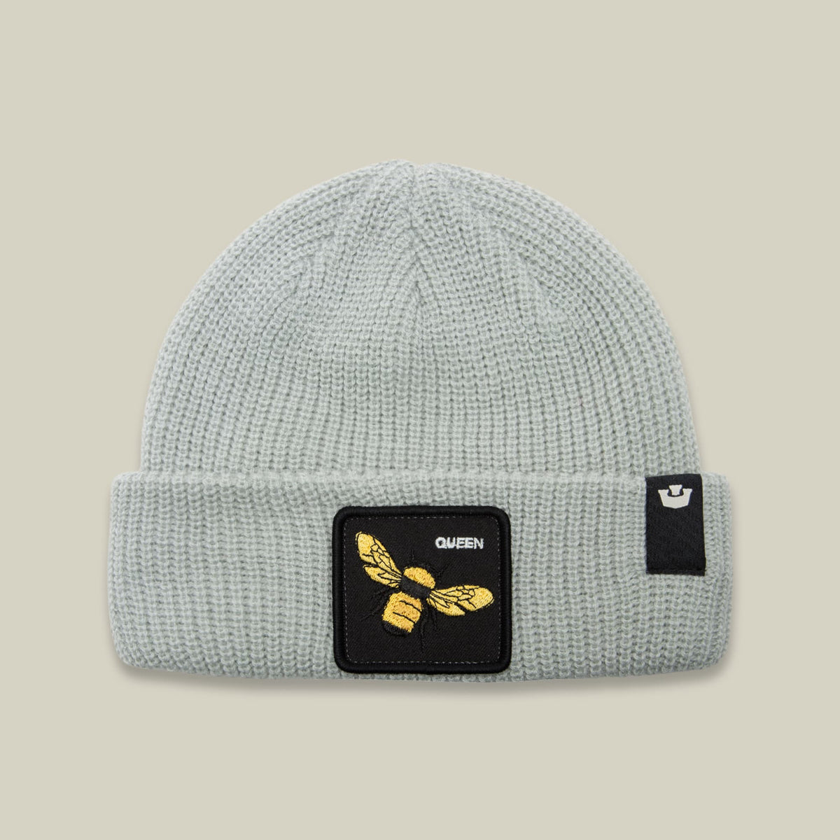 The Queen Bee Beanie by Goorin Bros is a light gray ribbed knit beanie featuring a black square patch with gold embroidered bee and "QUEEN" text on the front, displayed against a neutral background.