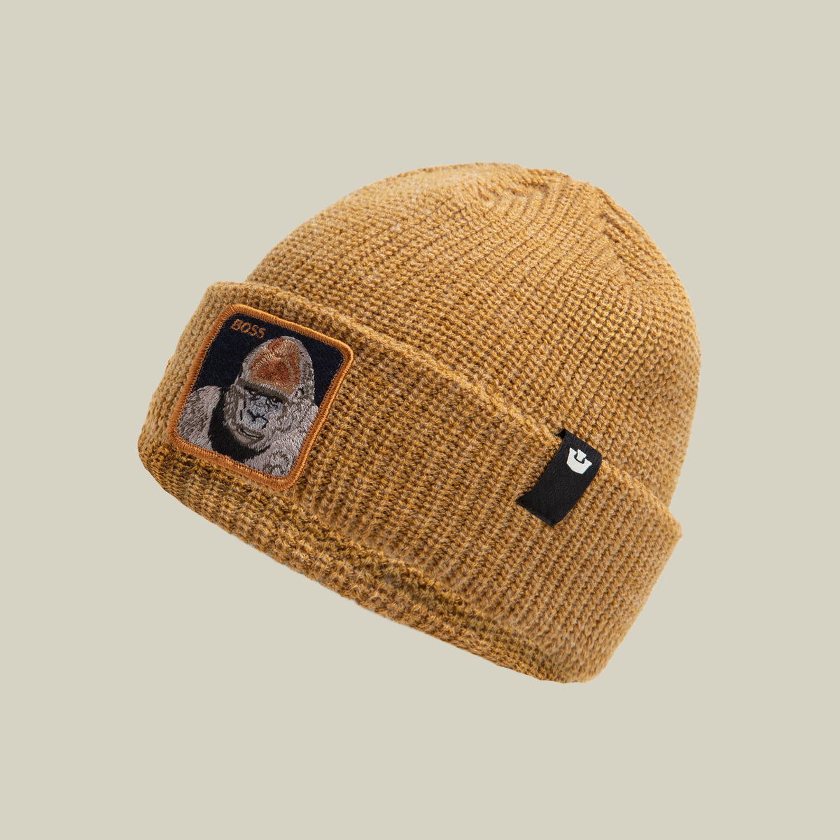 The Boss G Beanie by Goorin Bros is a tan knit beanie with a woven texture, featuring an embroidered portrait patch with orange trim on the front and a small black brand label on the side.