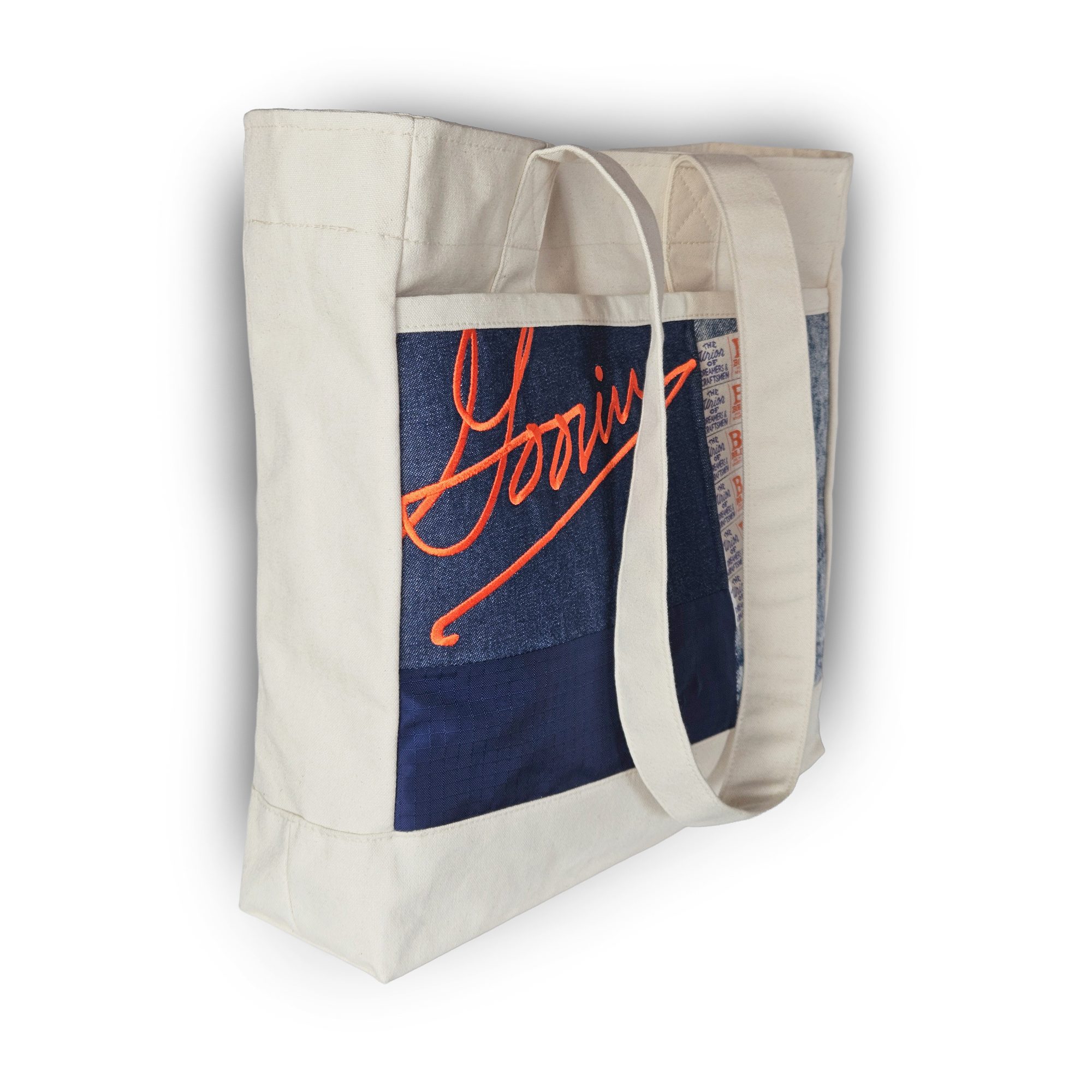 The Goorin Rework Tote by Goorin Bros. is a white canvas tote with long straps, featuring a front patchwork of upcycled blue and orange "Yooniv" text. Made in the USA.