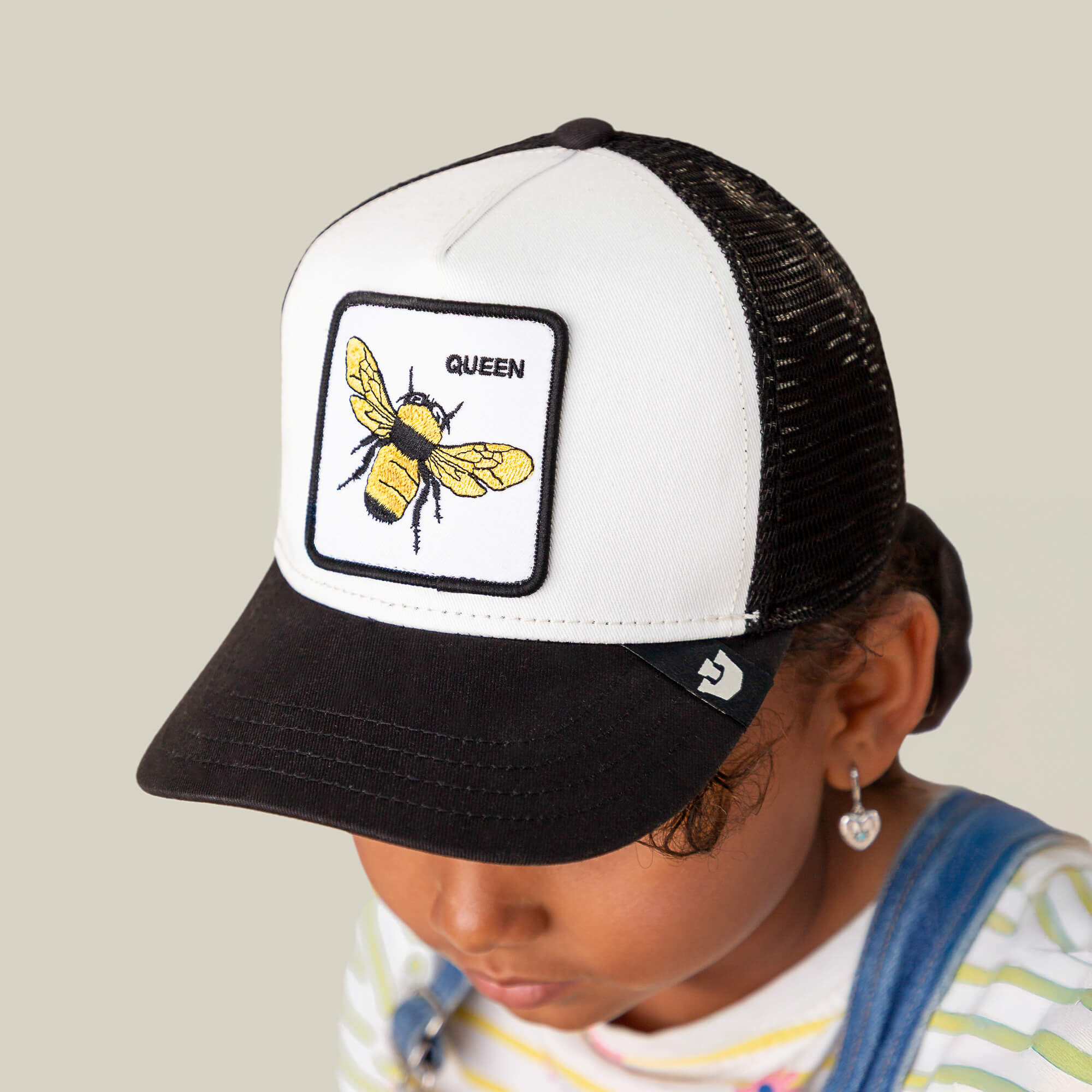 A child wears The Queen Bee Mini kids cap by Goorin Bros., a black and white hat with a golden bee patch labeled QUEEN on the front.