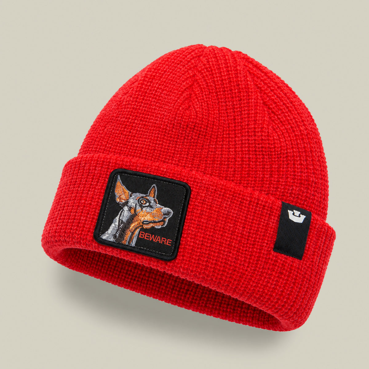 The Beware Dog Beanie is a bright red heavyweight knit beanie with a square patch featuring an embroidered Doberman head and the text BEWARE.