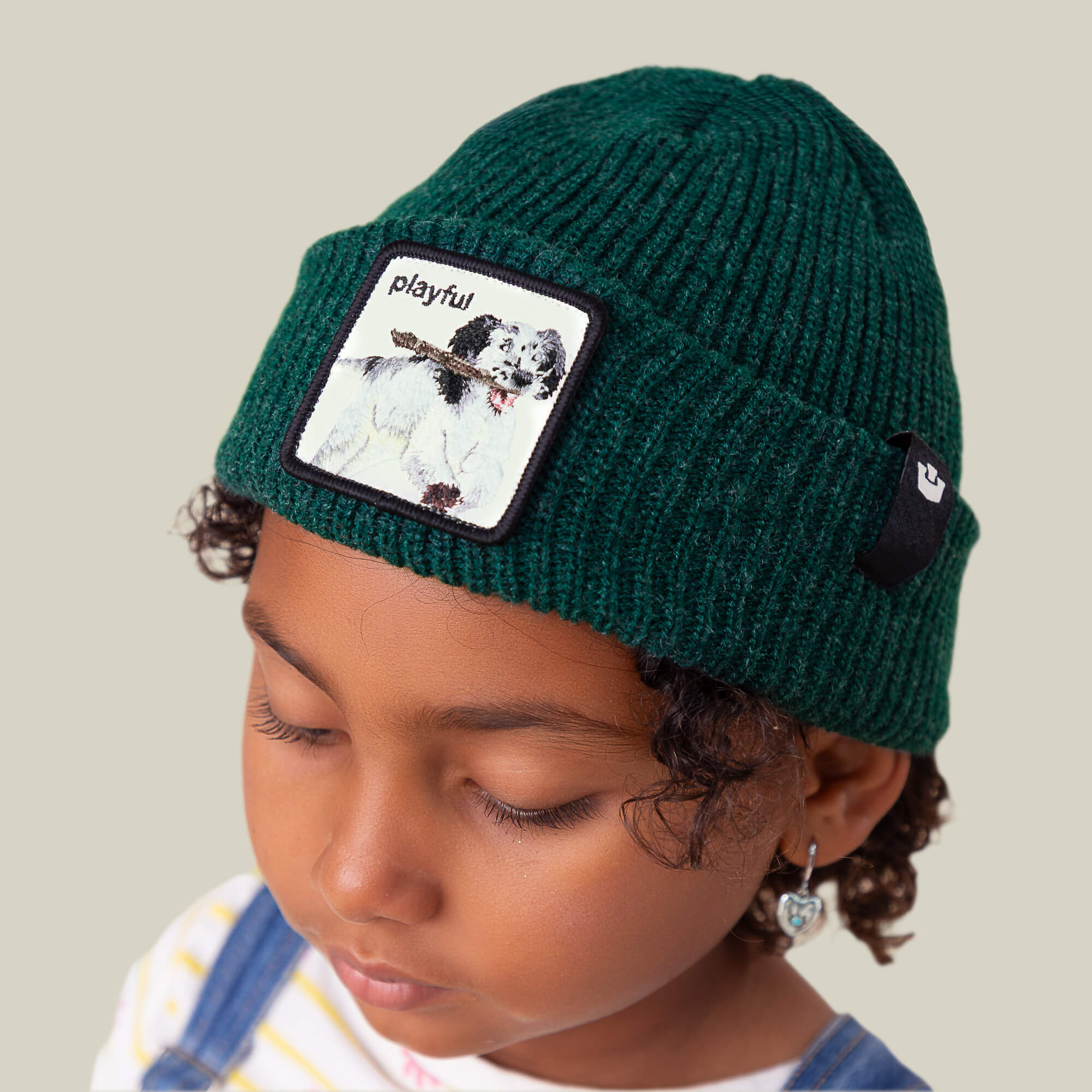 A young child wears The Playful Beanie Mini by Goorin Bros., a green kids hat featuring a playful dog patch and the word "playful," while looking down with a neutral expression.