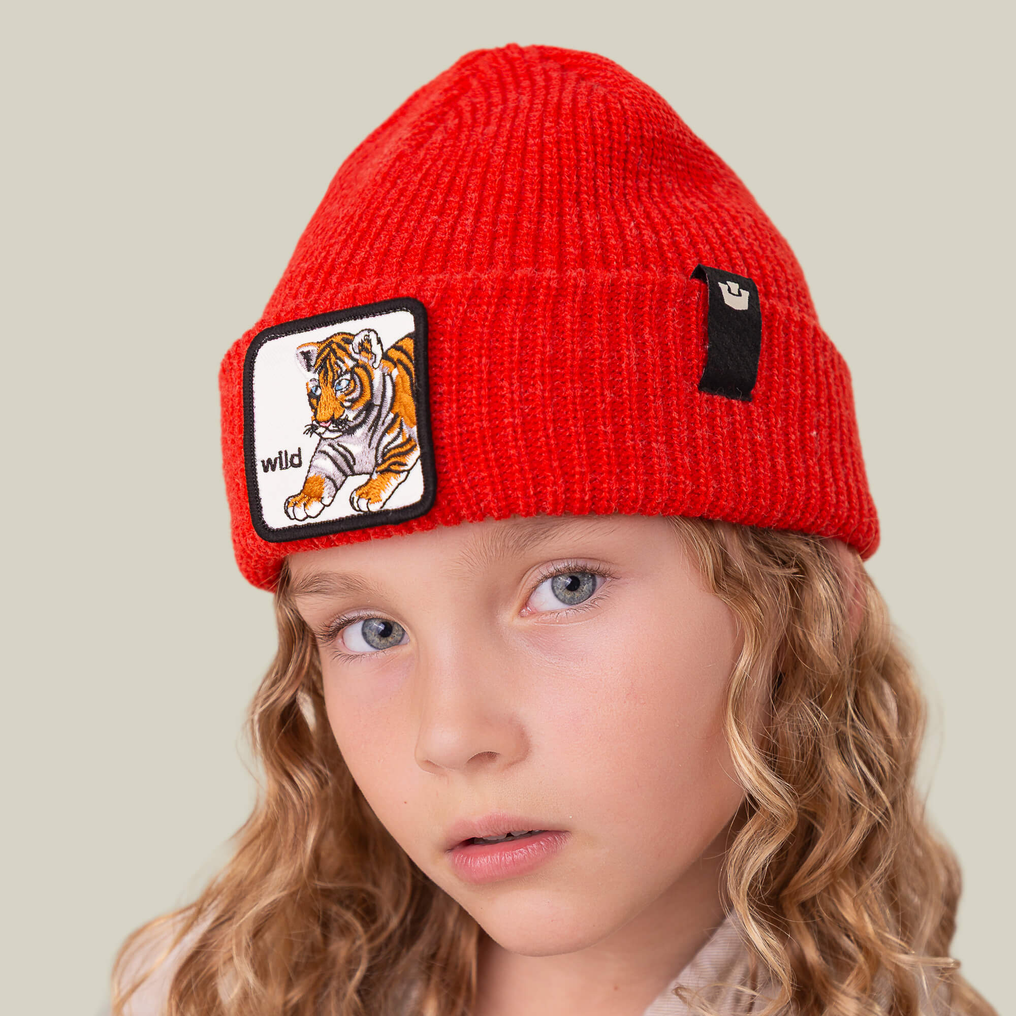 A child models The Wild Beanie Mini by Goorin Bros.—a bright red knit beanie with a tiger patch labeled "wild" and a small black tag—looking straight at the camera against a neutral background for a stylish look.