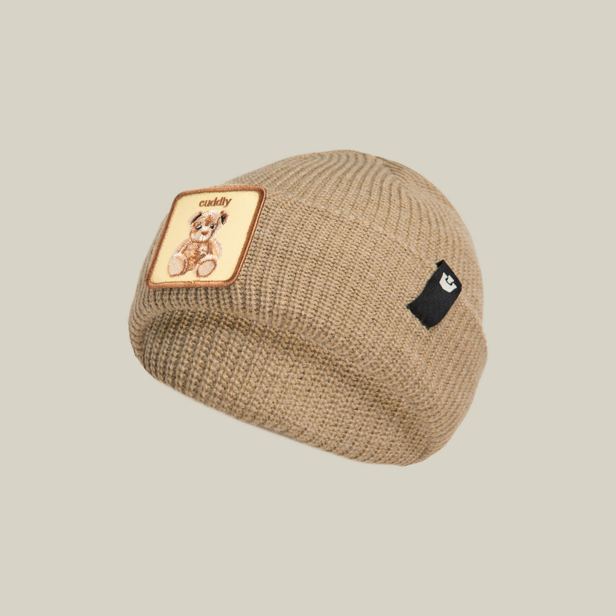 The Cuddly Beanie Mini by Goorin Bros. is a soft beige knit kids beanie featuring a square teddy patch with "cuddly" and a small black side tag.
