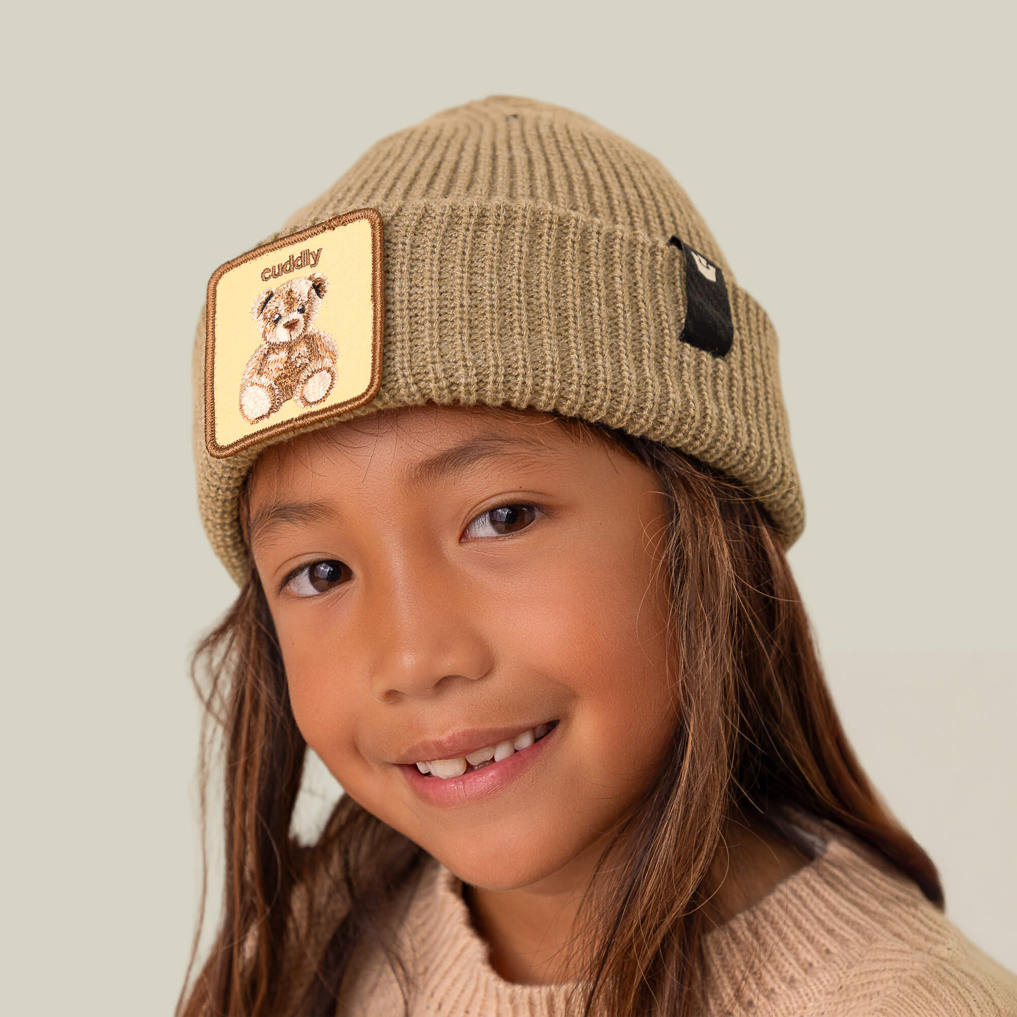 A smiling child wears The Cuddly Beanie Mini by Goorin Bros., featuring a teddy patch, paired with a beige sweater against a plain background.