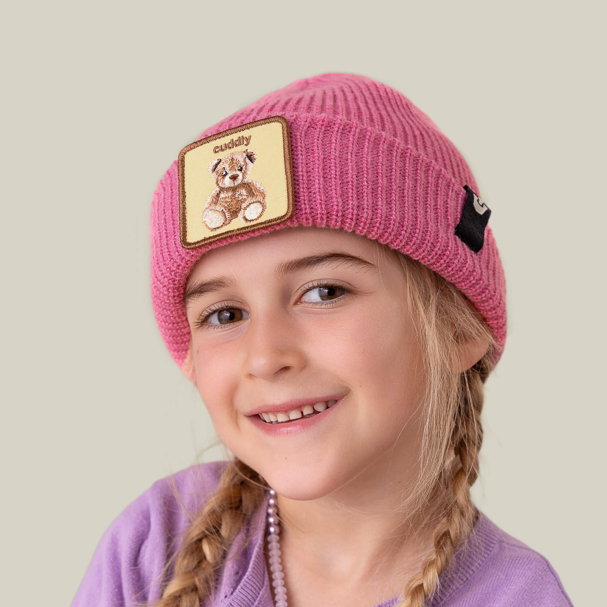 A young girl with blonde braids smiles, wearing Goorin Bros.' The Cuddly Beanie Mini—a pink kids' beanie featuring a teddy patch—and a purple top, against a plain background.