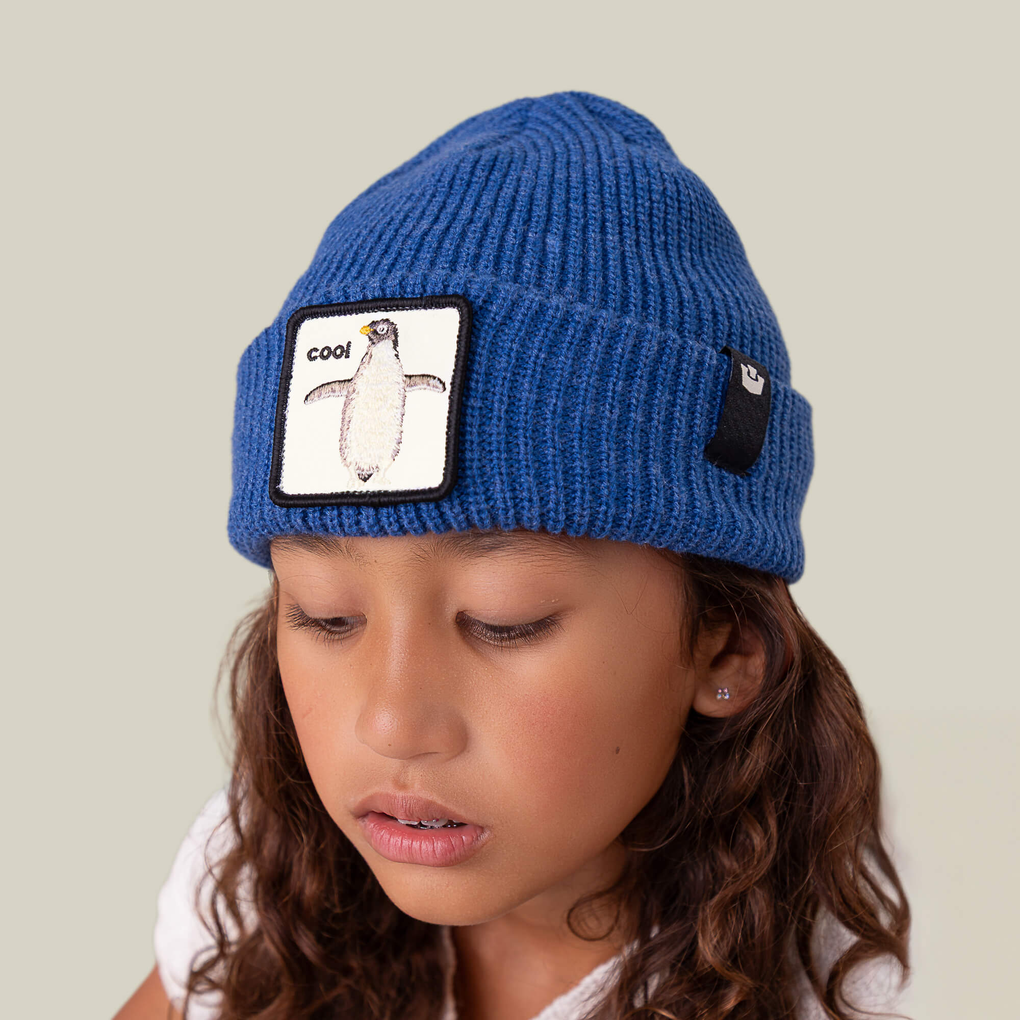A young girl wearing The Penguin Beanie Mini by Goorin Bros., featuring a "cool" penguin patch, looks down with a neutral expression.