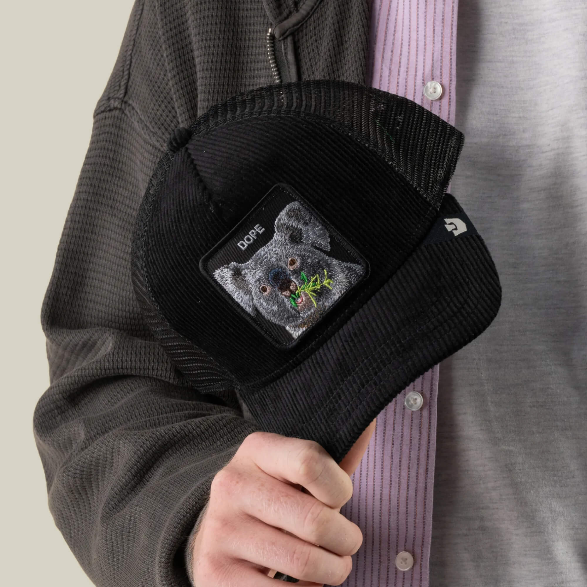 A person in a grey jacket and pink shirt holds the Goorin Bros. Corduroy Dope hat, a black corduroy trucker cap featuring an embroidered patch with a koala and the word “DOPE” on it.