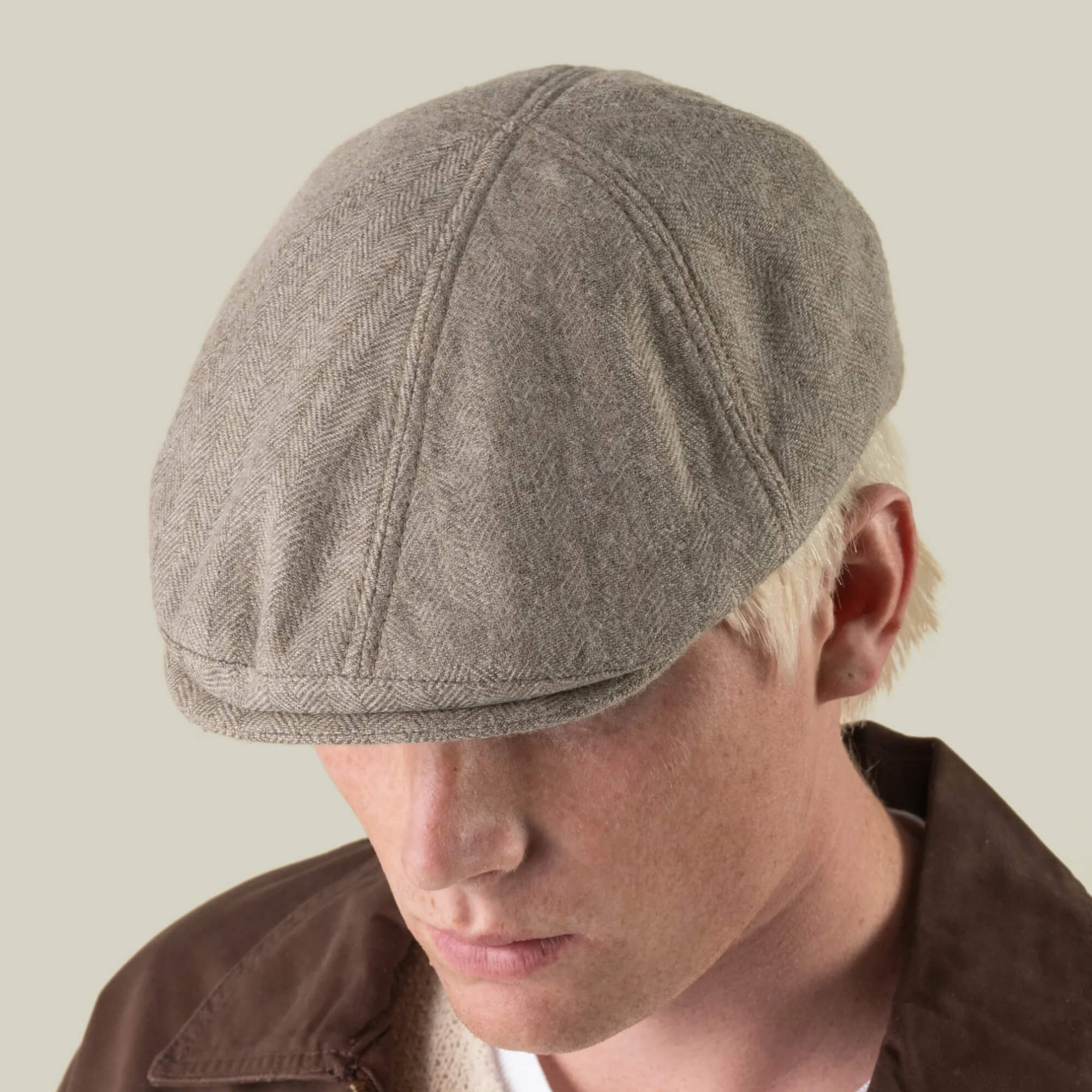 A person wearing the Goorin Bros. “Harry Bones” light brown herringbone flat cap, crafted from 100% wool, along with a brown jacket, looking downward against a plain background.