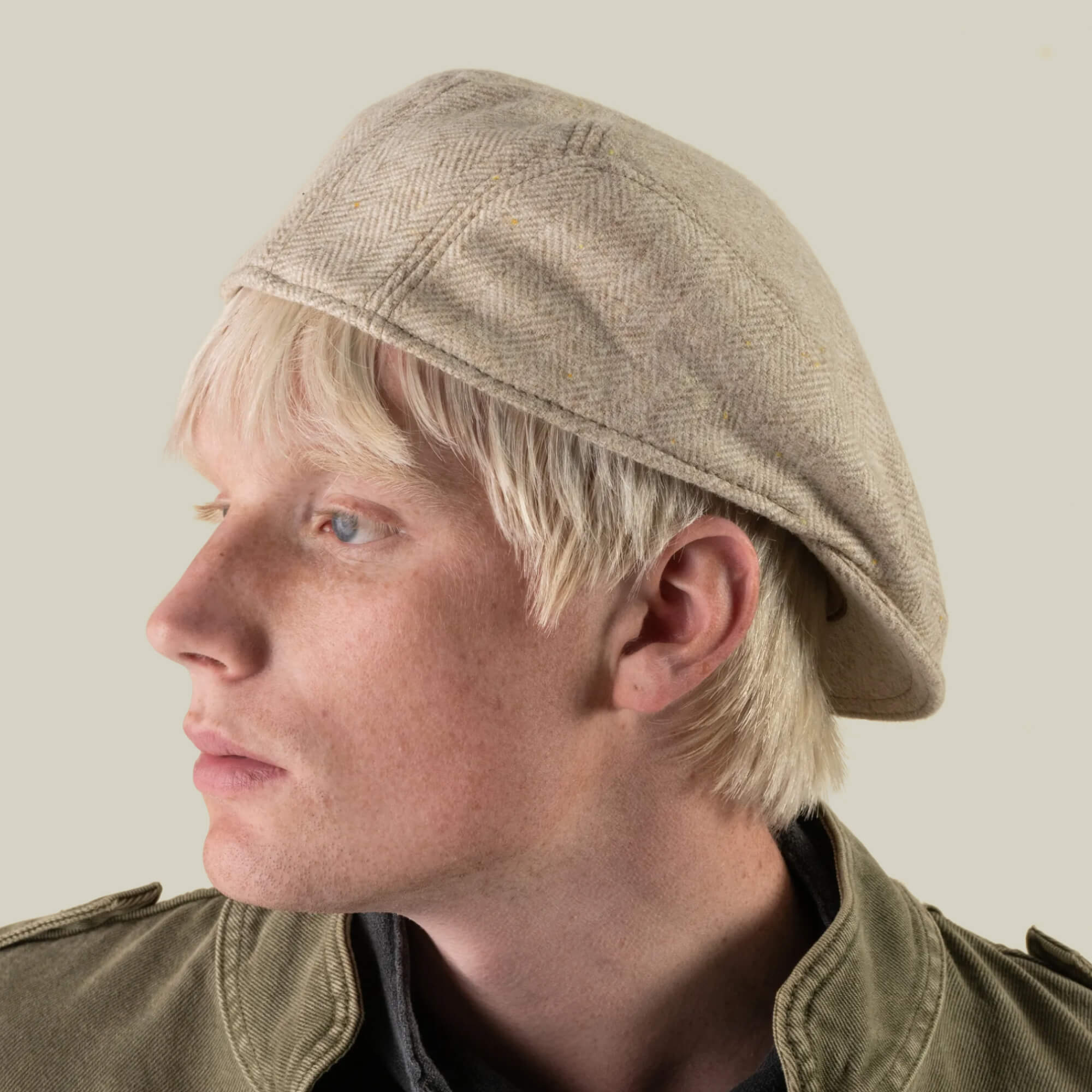 A person with light blond hair wears the Iris Fields flat cap by Goorin Bros.—a chalk cream headwear piece, hand-cut in the USA from 100% wool—as they look to the left. They are dressed in a green jacket against a plain background.