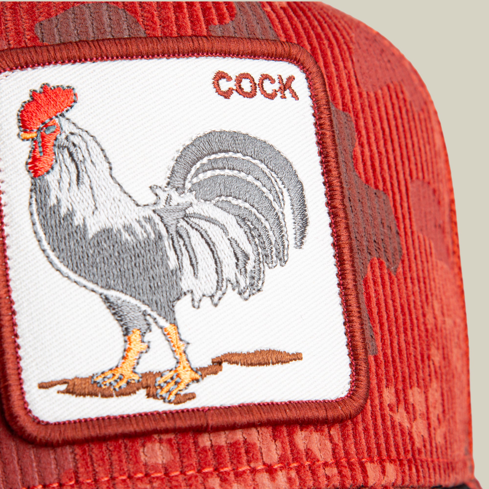 The Goorin Bros. Rooster Camo hat is a red corduroy trucker cap featuring a white patch with an embroidered rooster and the word “COCK” in red text.