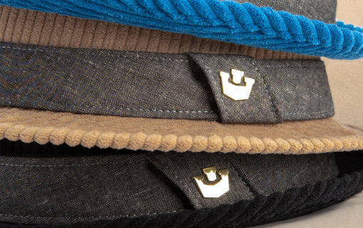 Close-up of three stacked hats with different colored brims—blue, beige, and black—each featuring a small metallic crown emblem on the band, showcasing Goorin's bold hat designs.