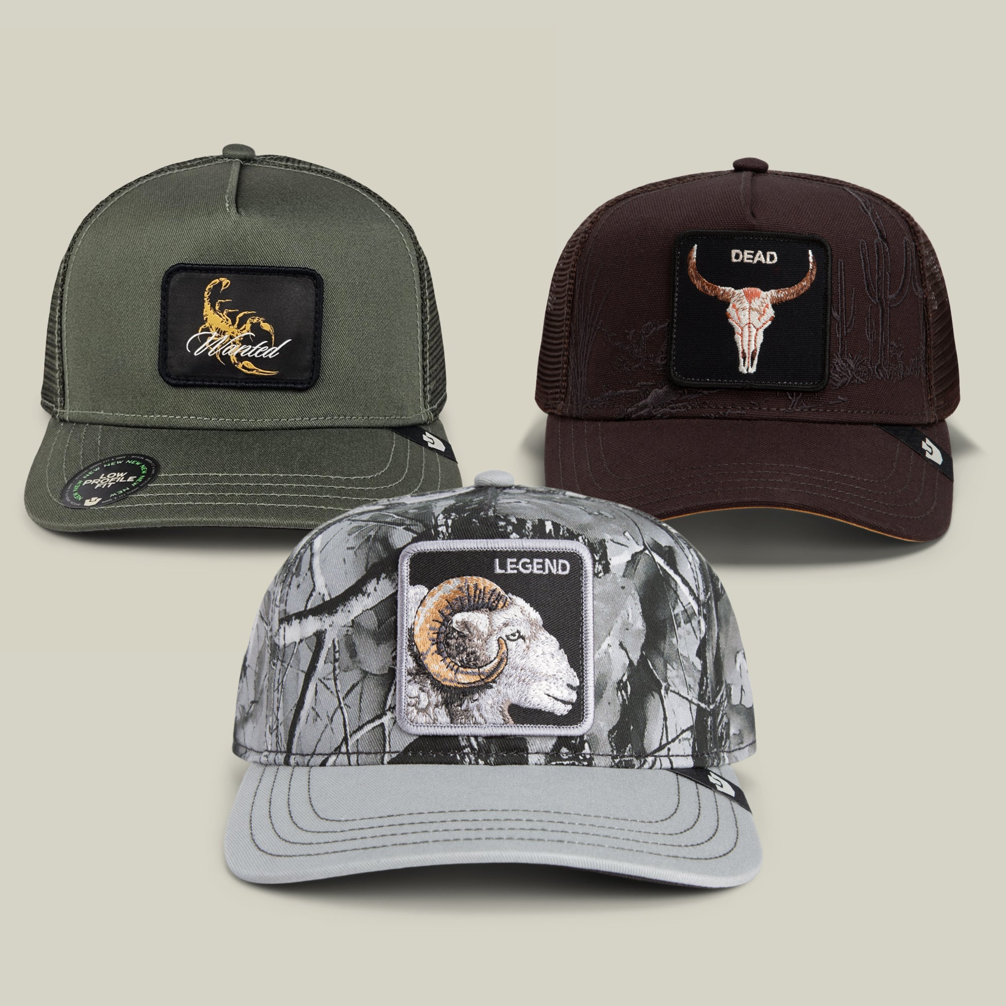 Three trucker hats from the Goorin Bros. Low Pro Bundle are displayed: an olive “Scorpion” hat with an eagle patch, a brown option featuring a maroon “Longhorn Skull DEAD” patch, and a legend grey camo hat with a “Ram LEGEND” patch.