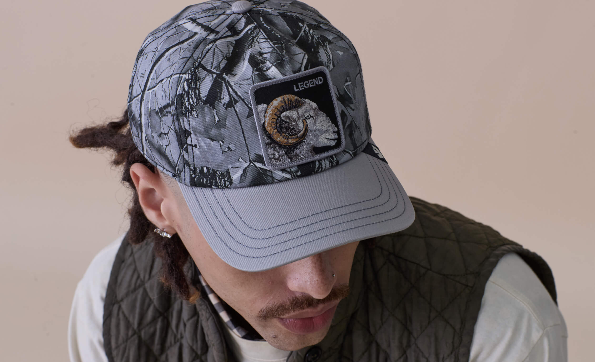 A man in a quilted jacket and a camouflage-patterned cap with a ram patch labeled Legend looks downward, showcasing bold hat designs and American made durability.