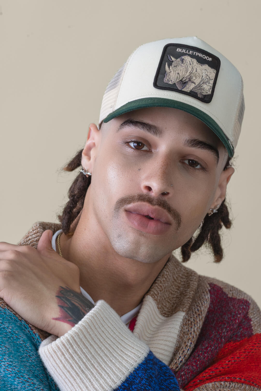 A person with a mustache and dreadlocks wears an American made rhino patch cap, hoop earrings, and a multicolored sweater, looking at the camera with a neutral expression.