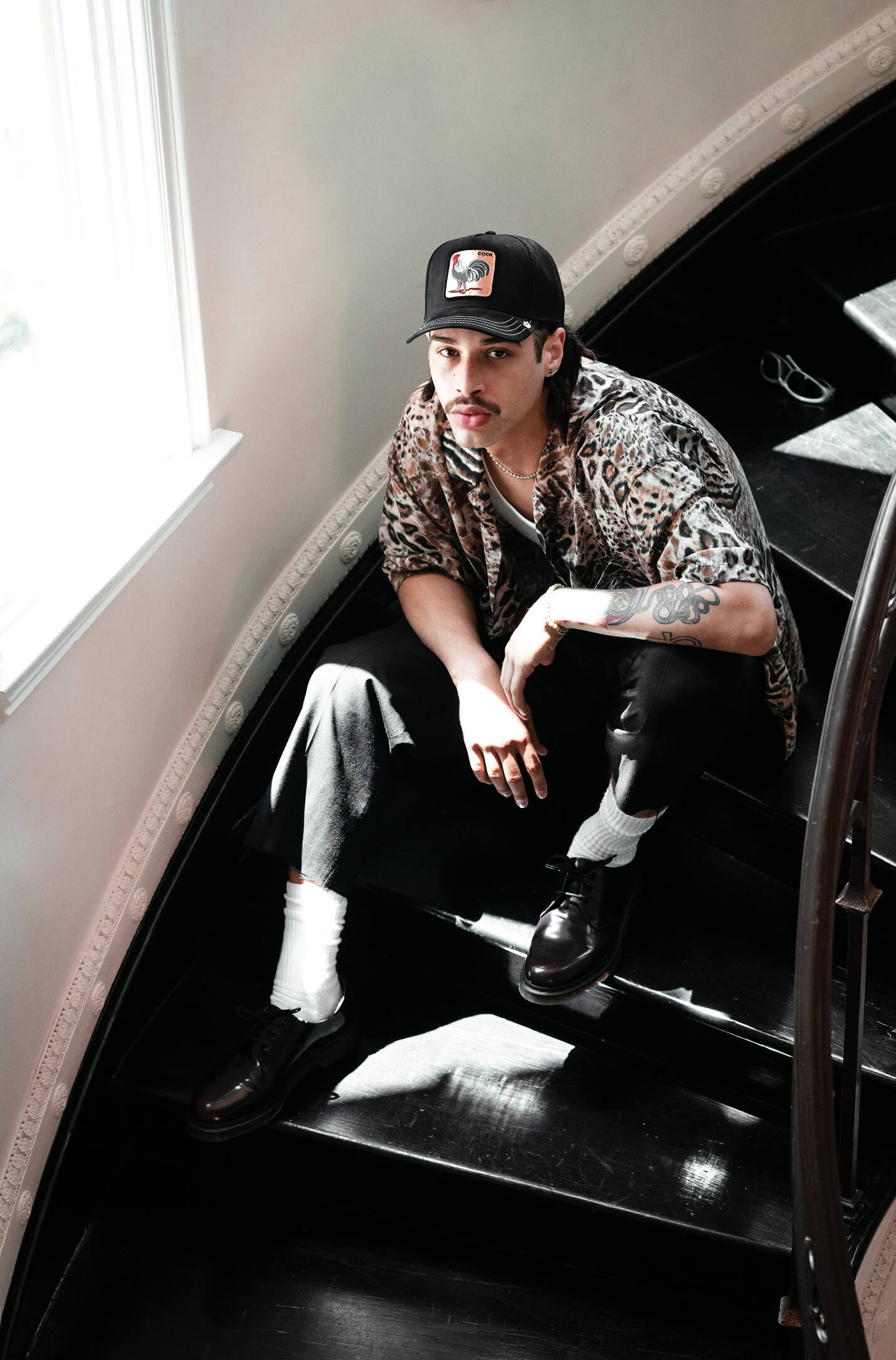A man wearing a patterned shirt, dark pants, and a Goorin fedora hat sits on a black staircase near a window, looking up at the camera with light streaming in.