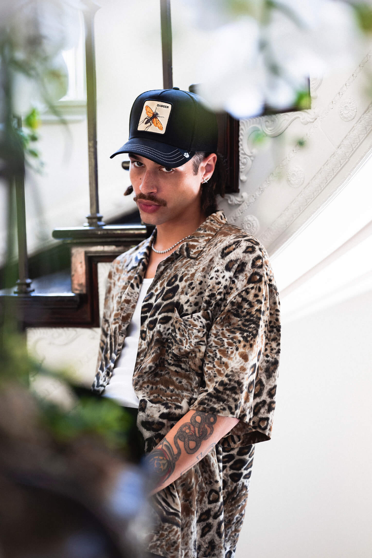 A man with a mustache, wearing a leopard print shirt, white tank top, beaded necklace, and a Goorin trucker hat with a butterfly patch, stands by a staircase with one arm tattooed.