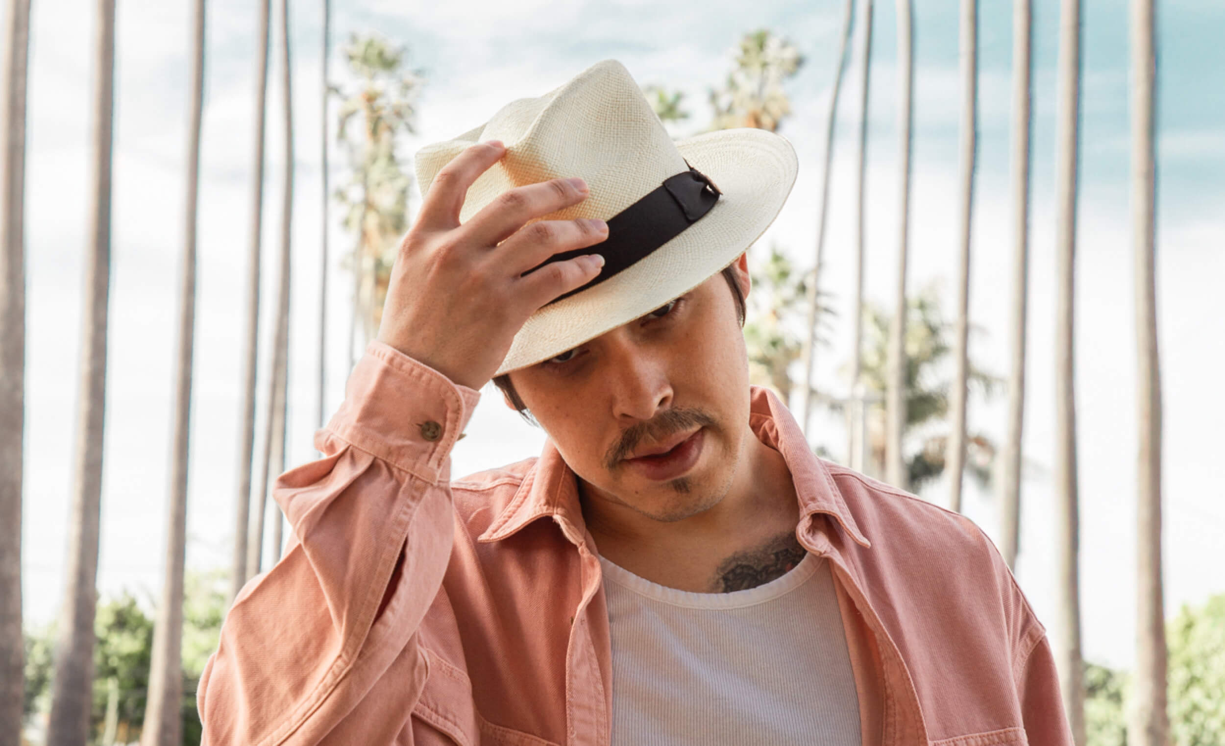 Man adjusting a white fedora hat with a black band against a blurred natural background