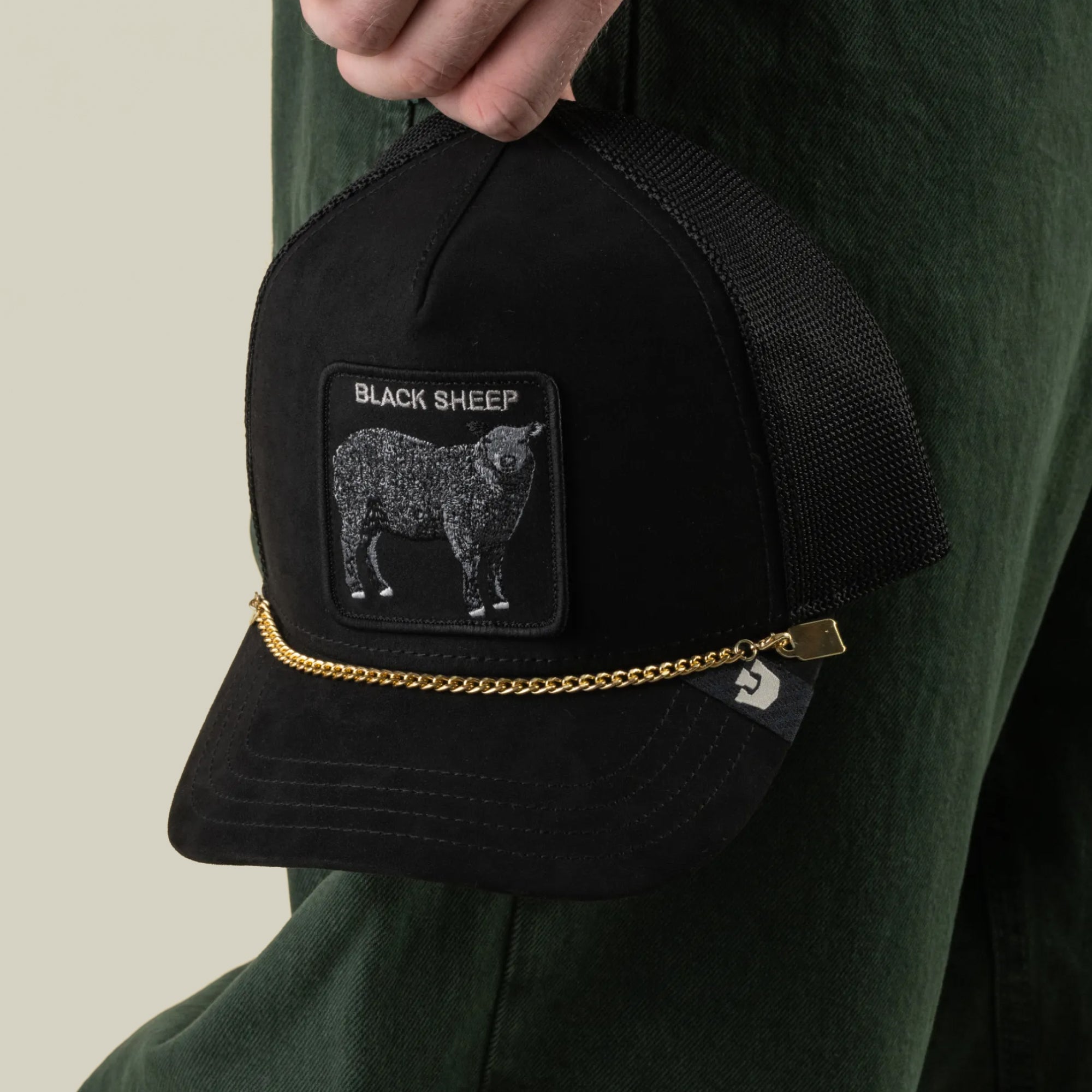 A hand holds a black mesh trucker hat by Goorin Bros., featuring a BLACK SHEEP patch, a sheep graphic, and the stylish Mini CapLoc Chain—hat jewelry with a gold chain and double clip chains adorning the front. The person is wearing dark green pants.