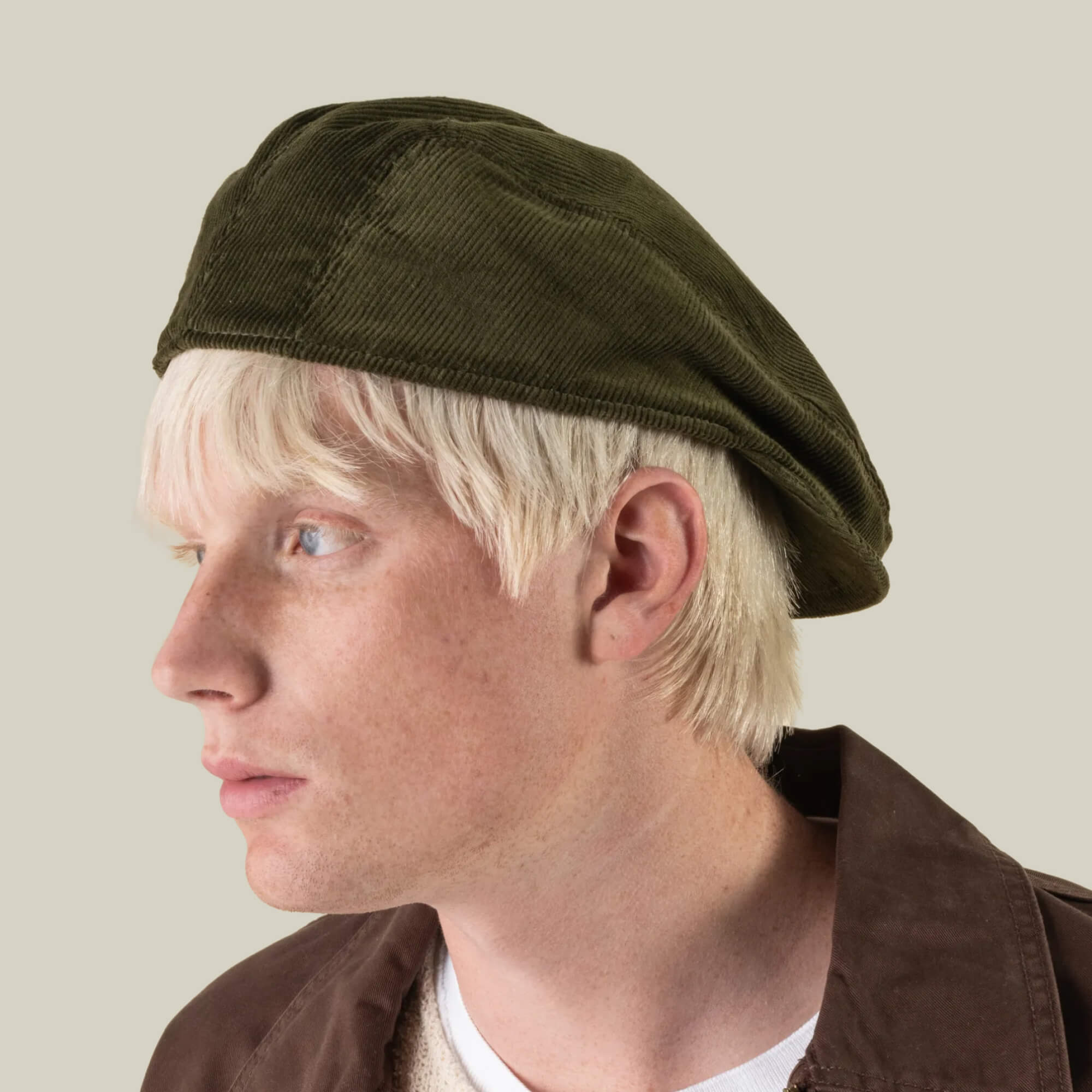 A person with light skin and blonde hair is wearing the Maxwell Park beret by Goorin Bros.—a durable, hand-cut corduroy hat in olive green, crafted in the USA—styled with a brown jacket and gazing to the left against a plain light background.