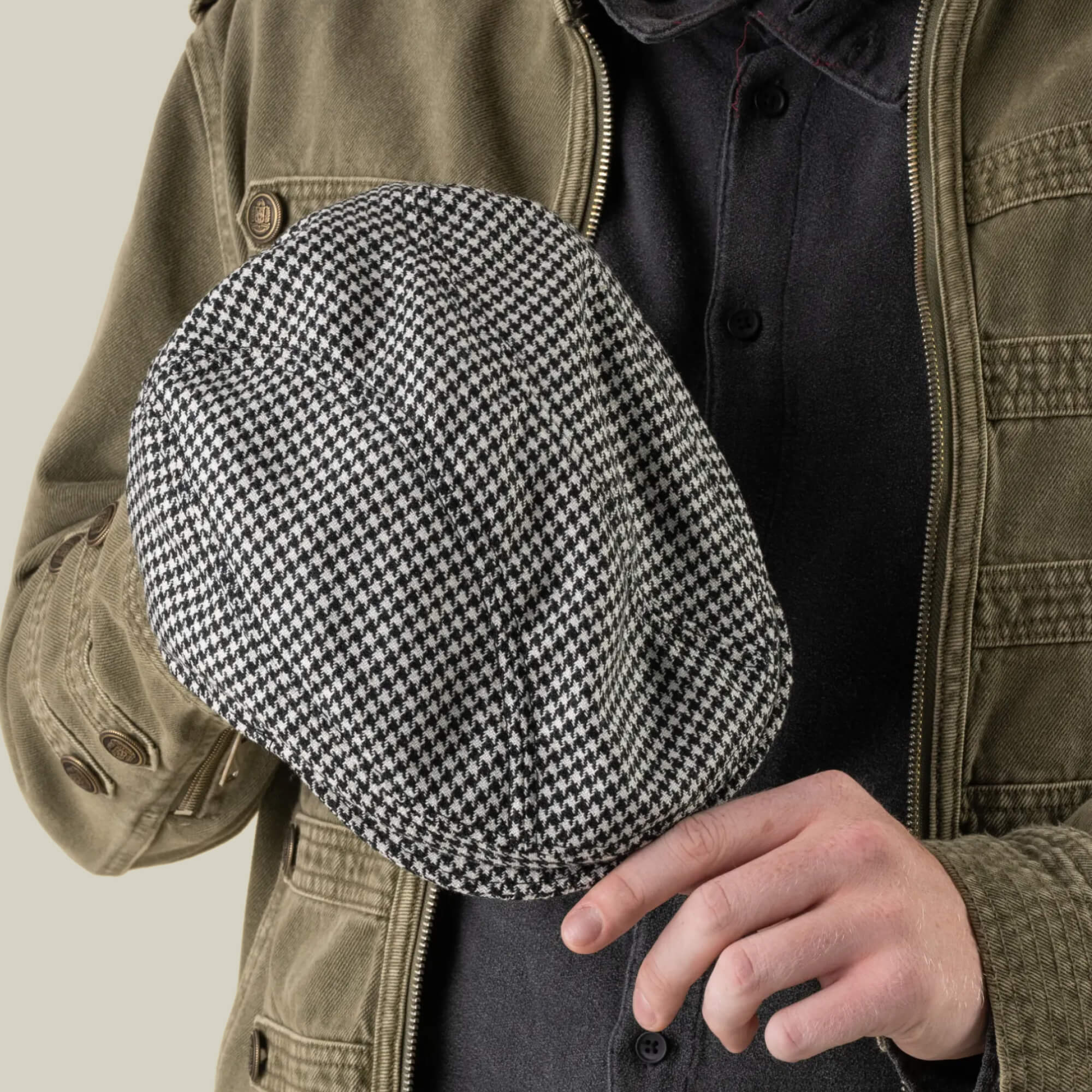 A person in a green jacket and dark shirt holds the Goorin Bros. Norwood black-and-white houndstooth wool flat cap in one hand and touches the brim with the other hand.