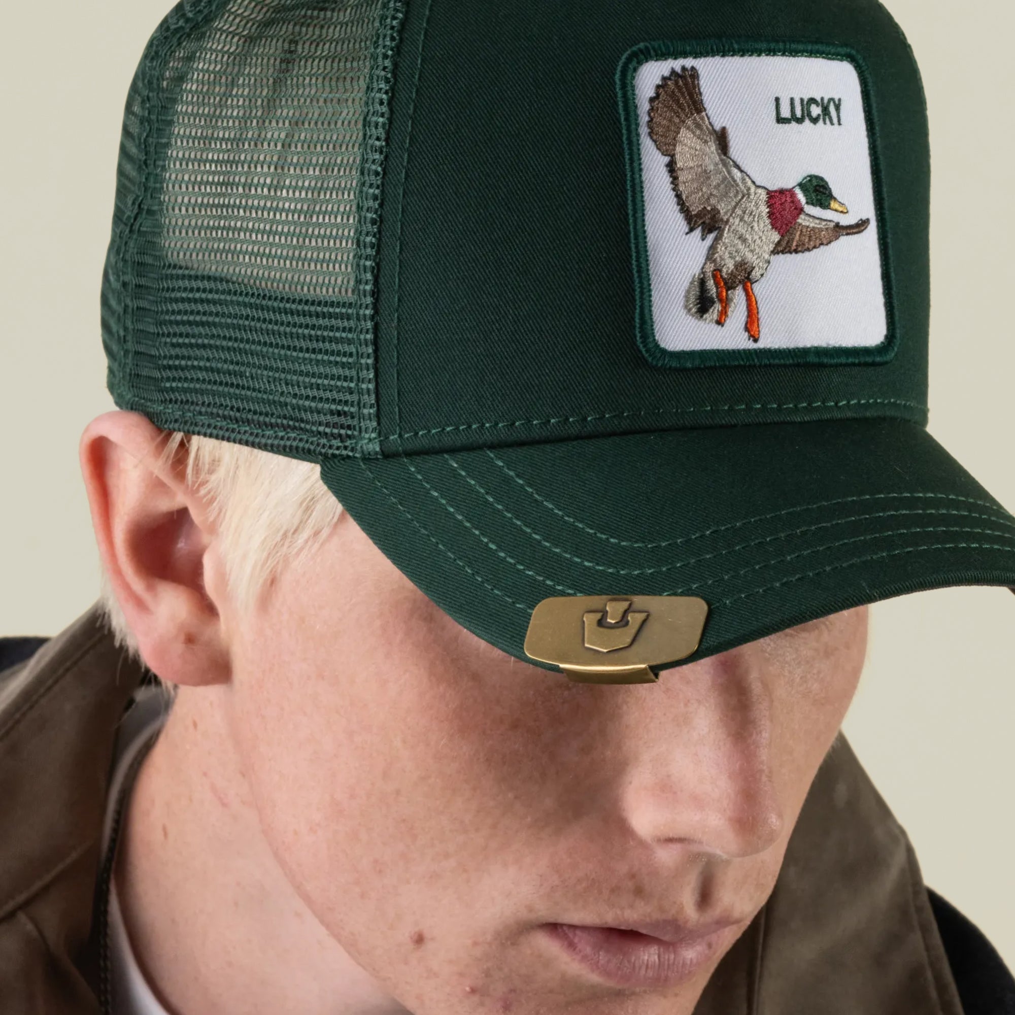 A person wearing a dark green mesh baseball cap with an embroidered patch of a flying duck and the word LUCKY, by Goorin Bros. The cap features the Castle CapLoc, a stylish zinc alloy Brim Clip accessory, attached to the brim.