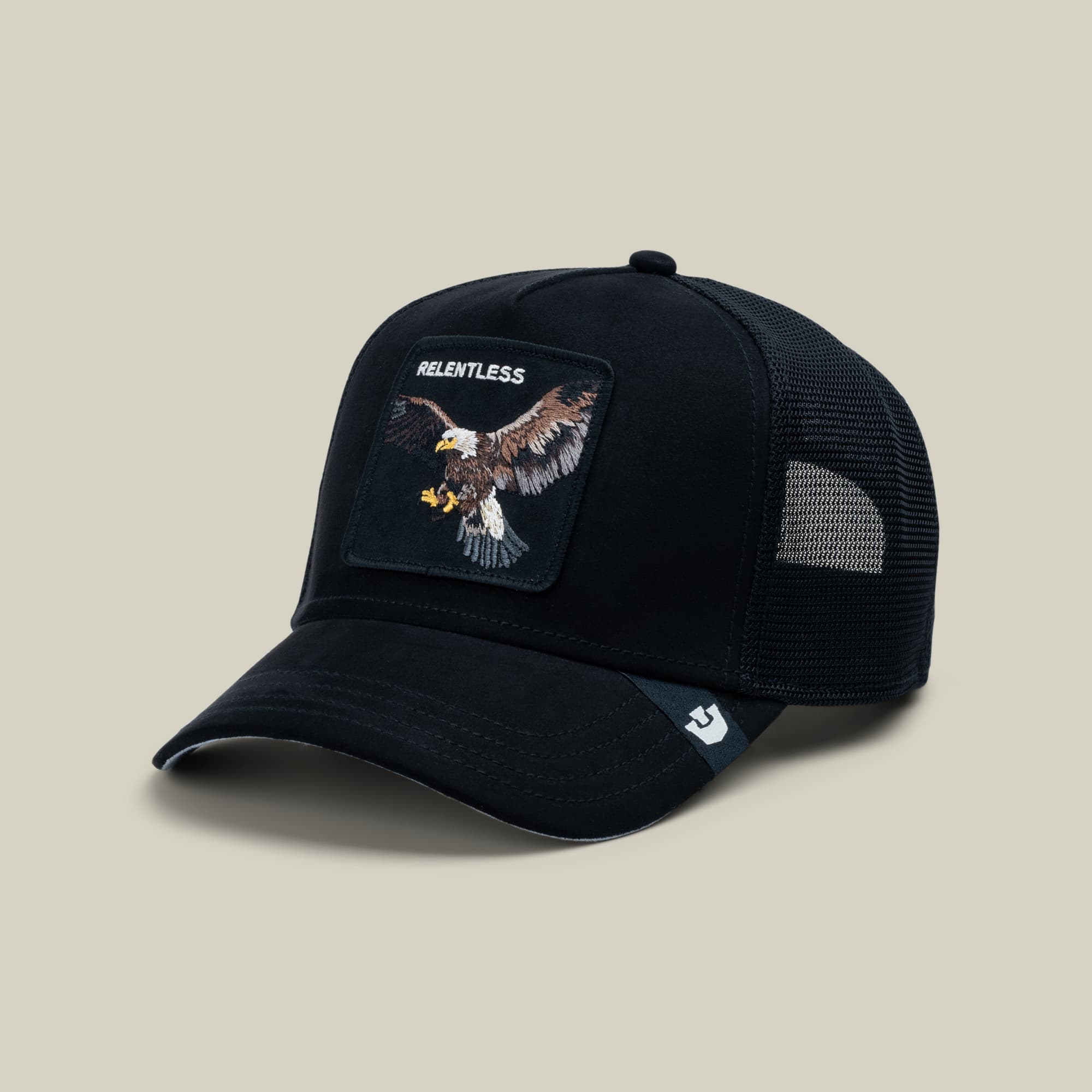 The Goorin Bros. Suede Relentless Eagle Trucker is a black mesh-back cap featuring an embroidered eagle patch with "Relentless" text on the front, displayed on a neutral beige studio backdrop.
