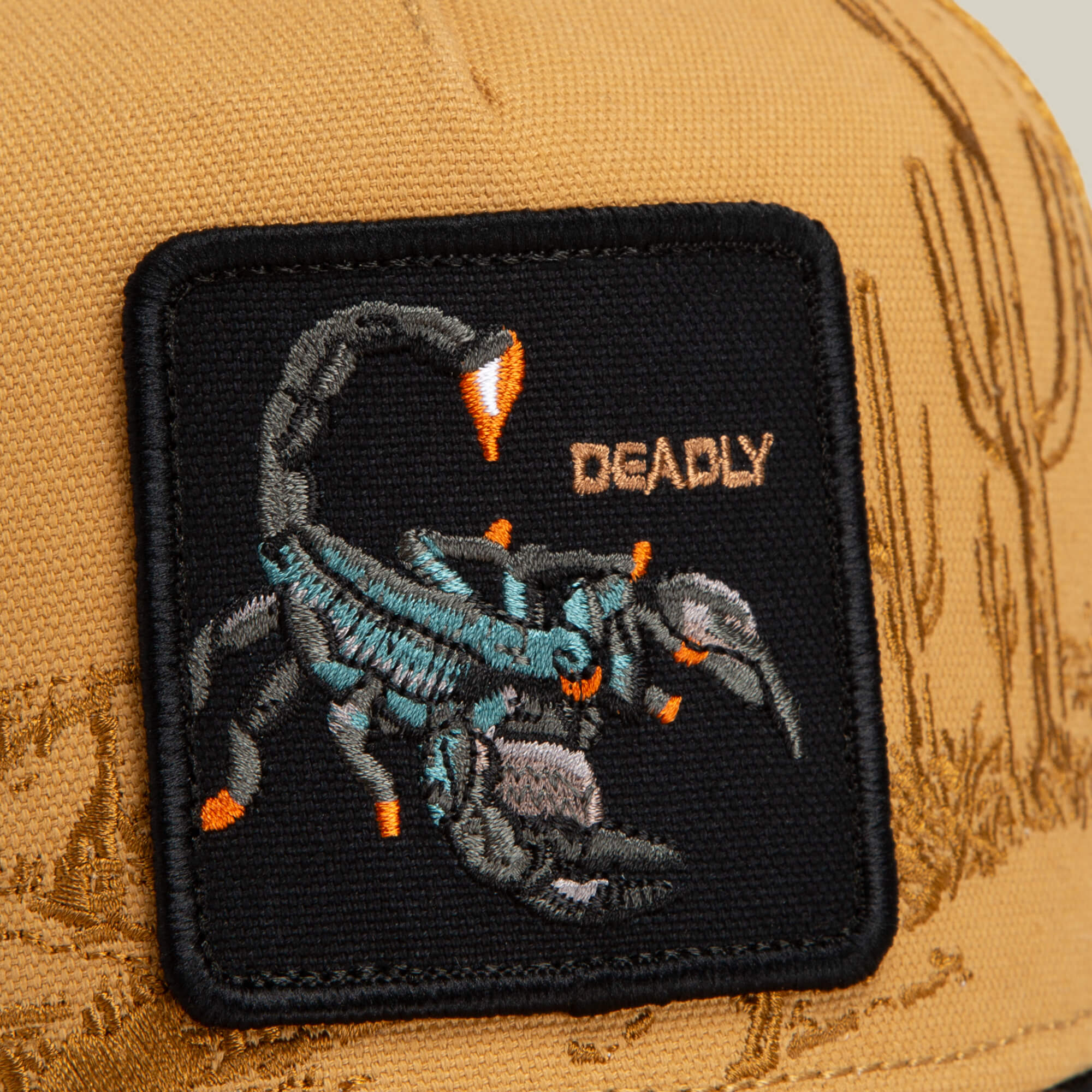 Close-up of the Goorin Bros. Badlands Scorpion tan cap, showcasing a black patch with an embroidered blue and gray scorpion and the word "DEADLY" in gold lettering. Desert-inspired stitching decorates the background.