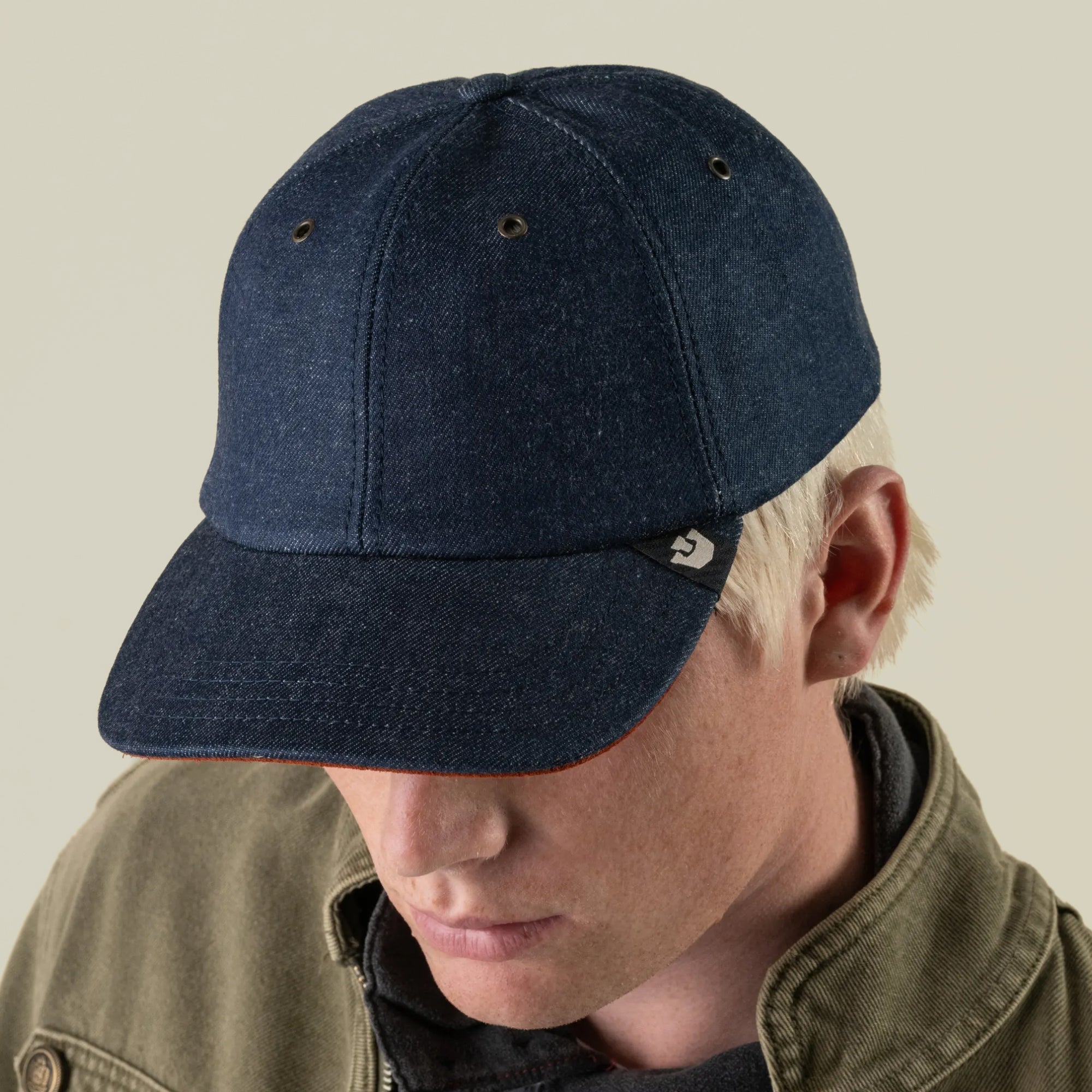A person with light hair wears the Goorin Bros. Legend Ridge dark denim baseball cap—a mixed-material accessory made in the USA—paired with an olive green jacket, looking downward against a plain background.