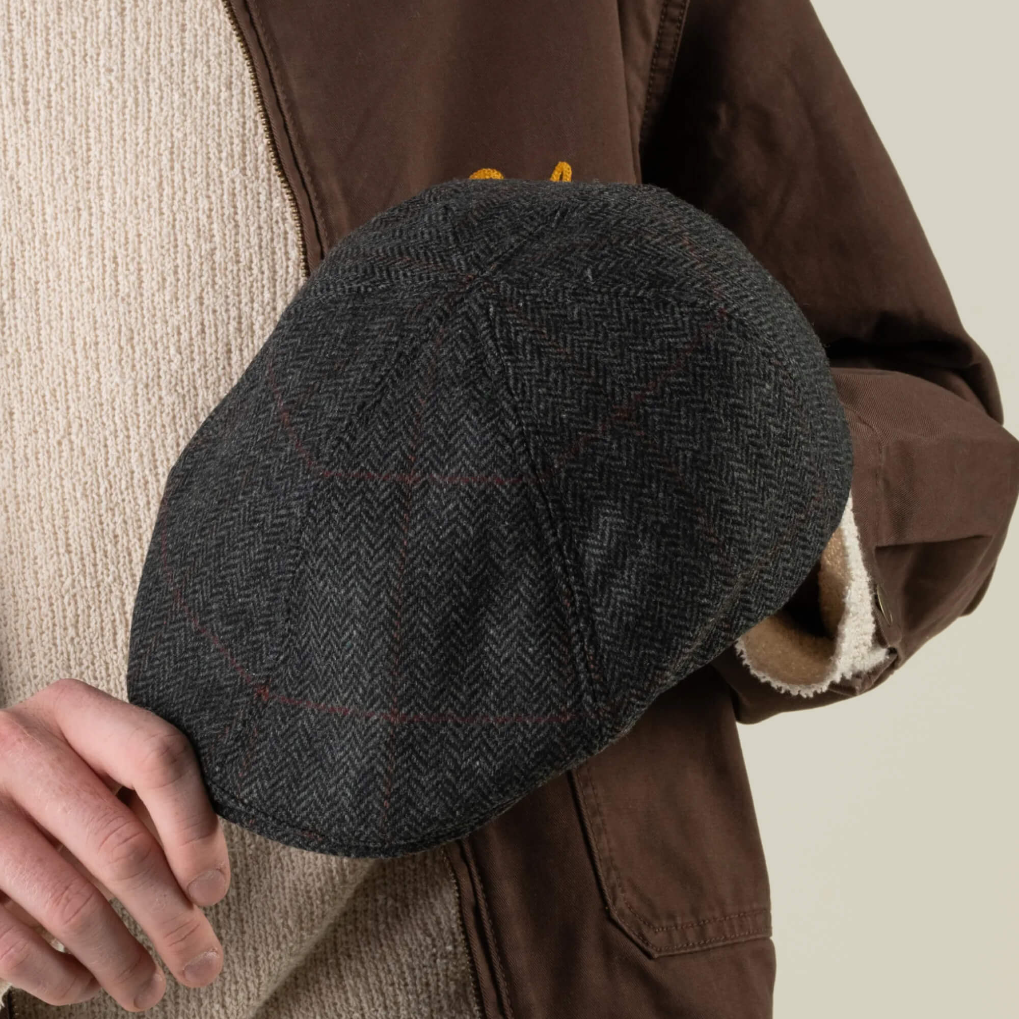 A person holds the Goorin Bros. Harry Bones flat cap—dark gray herringbone, crafted from 100% wool and made in the USA—while wearing a brown jacket and beige sweater against a neutral background.